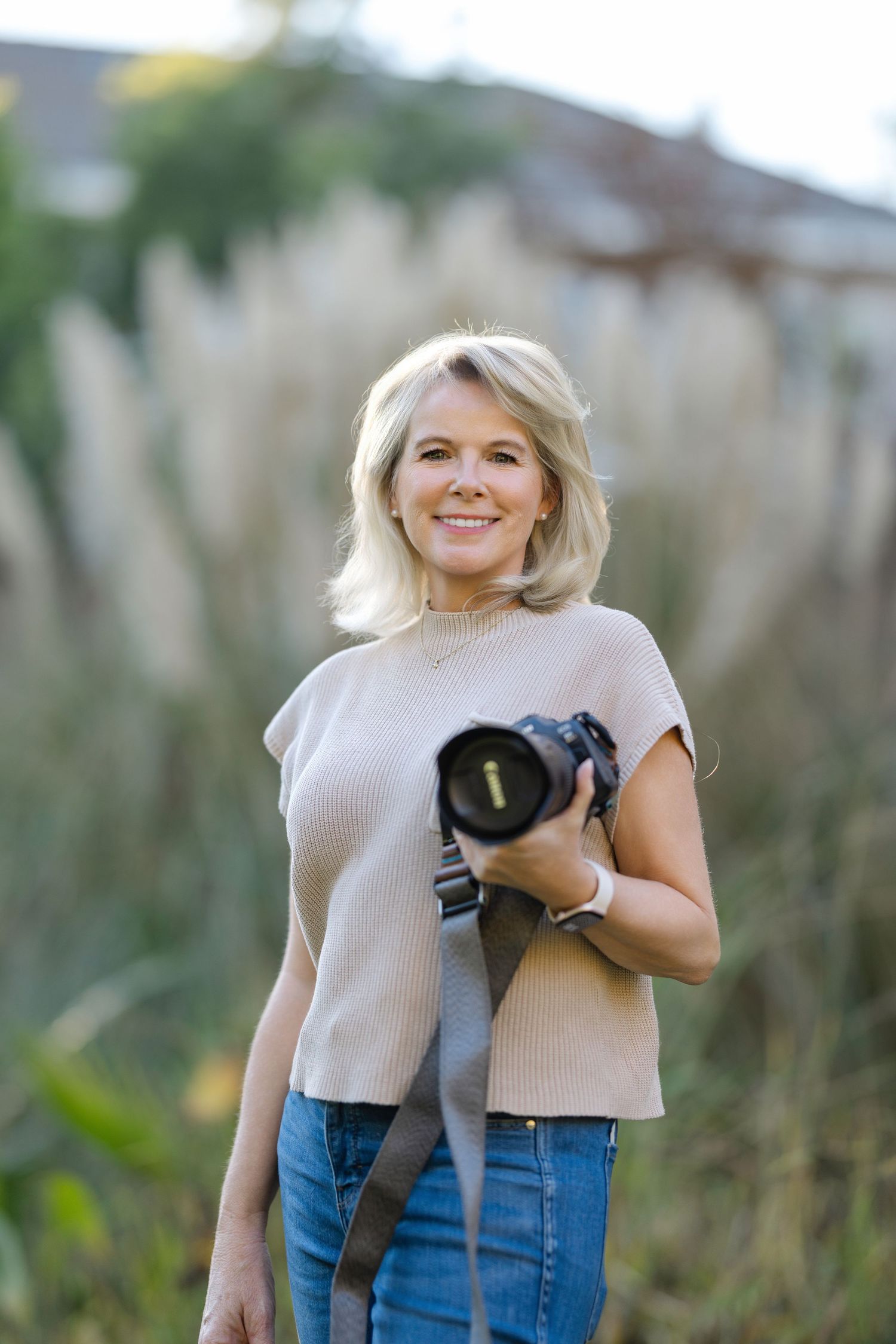 Photographer in casual attire smiling while holding professional camera equipment in natural setting.