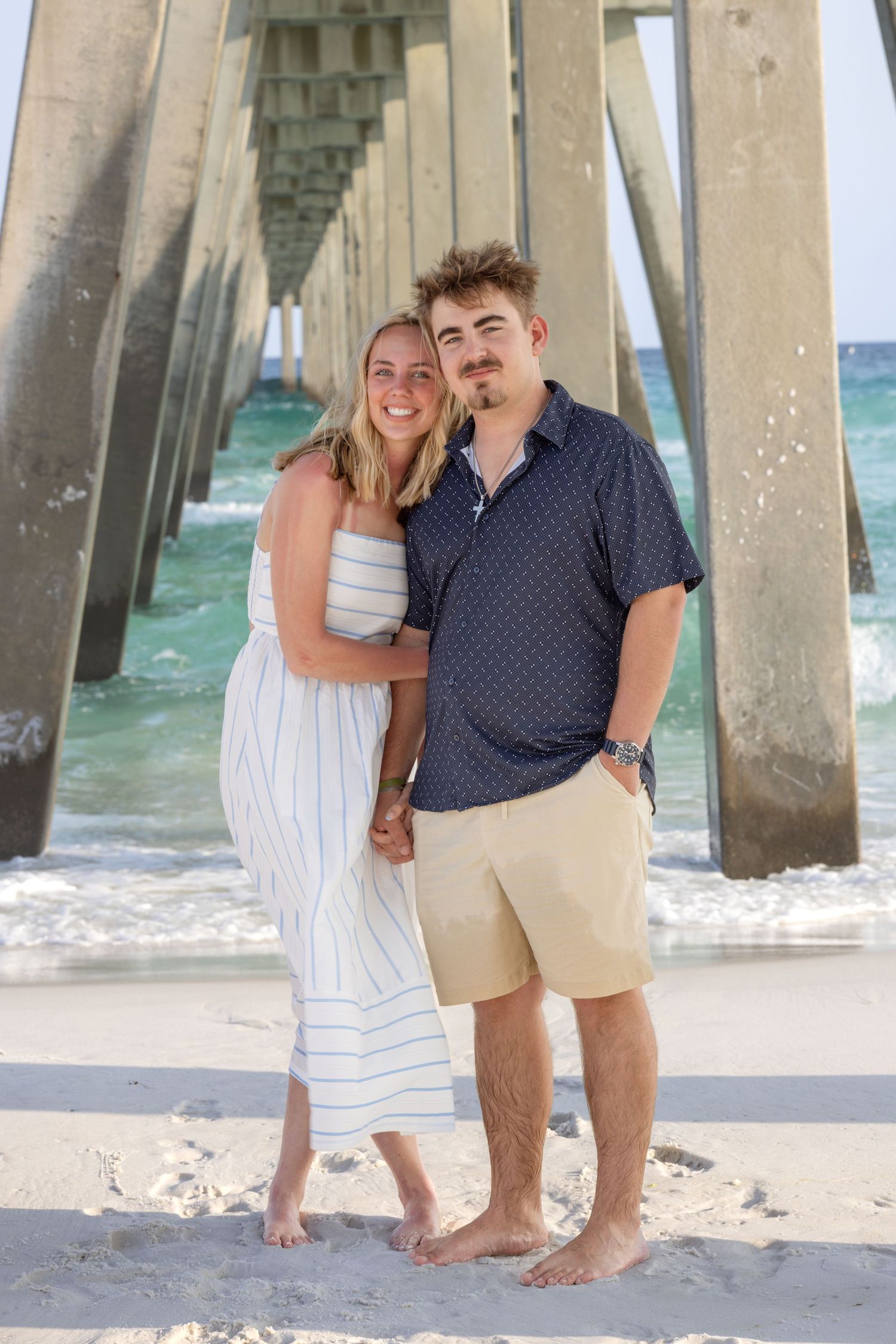 Happy couple posing barefoot under a beach pier, woman in striped sundress and man in navy shirt.