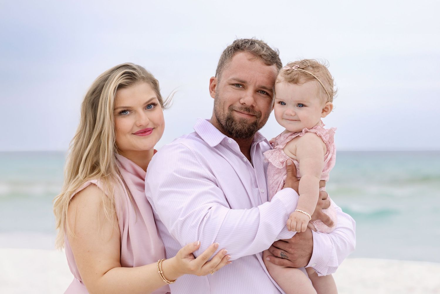 A smiling couple holding their baby girl poses together on a sandy beach with ocean waves in the background.