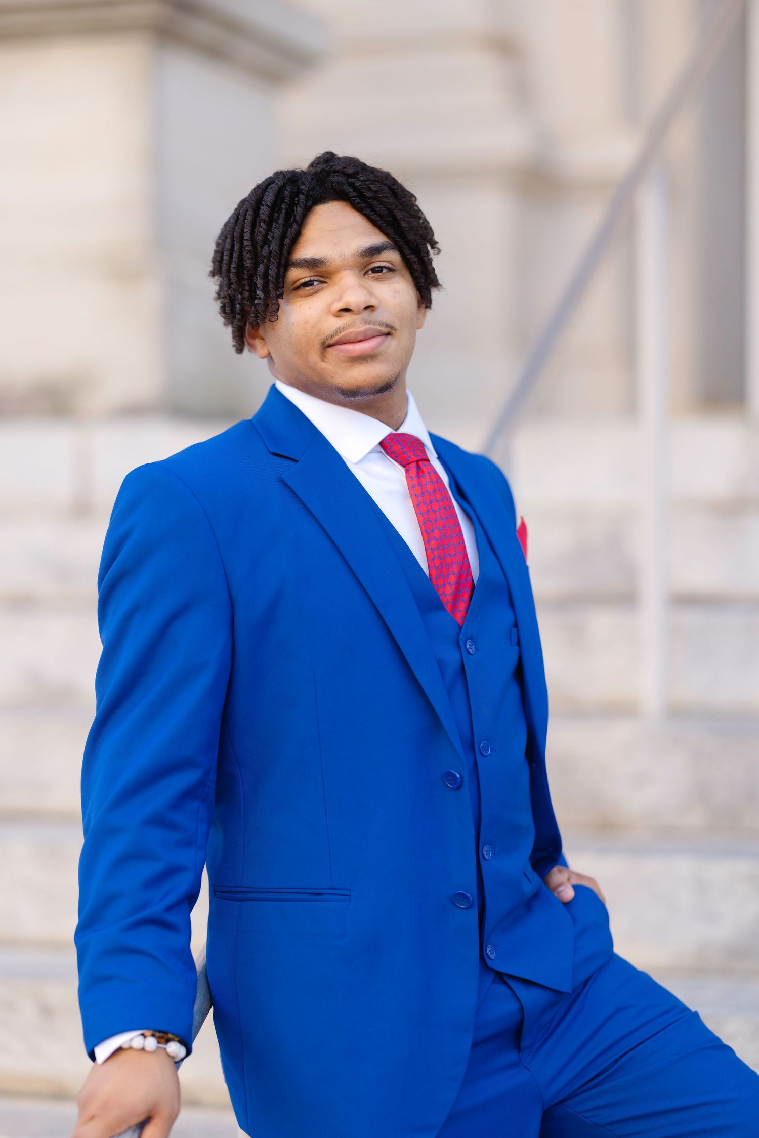 Young man in royal blue three-piece suit with red tie and pocket square, posing confidently outdoors.