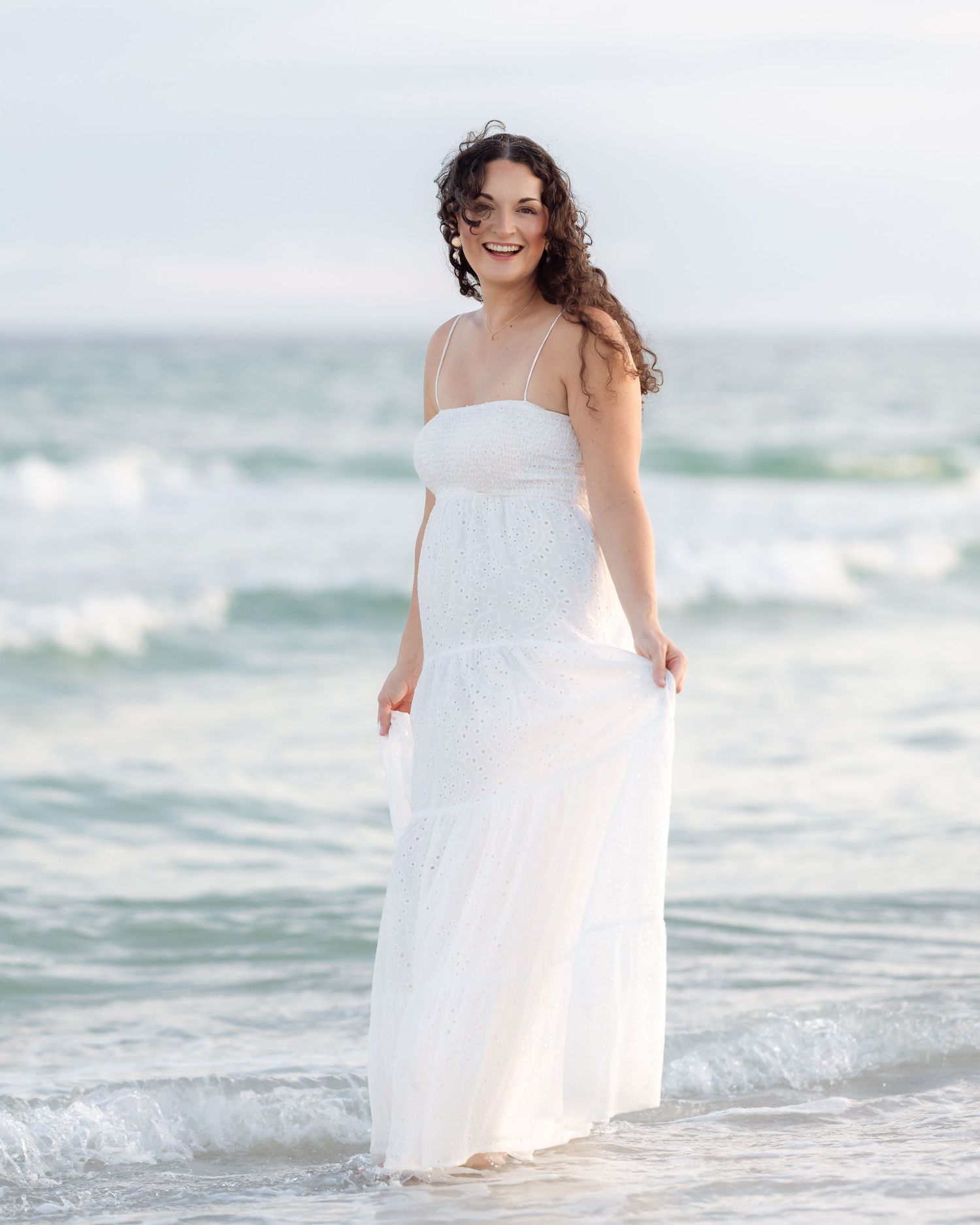 Smiling pregnant woman in elegant white maxi dress standing in ocean waves on beach.