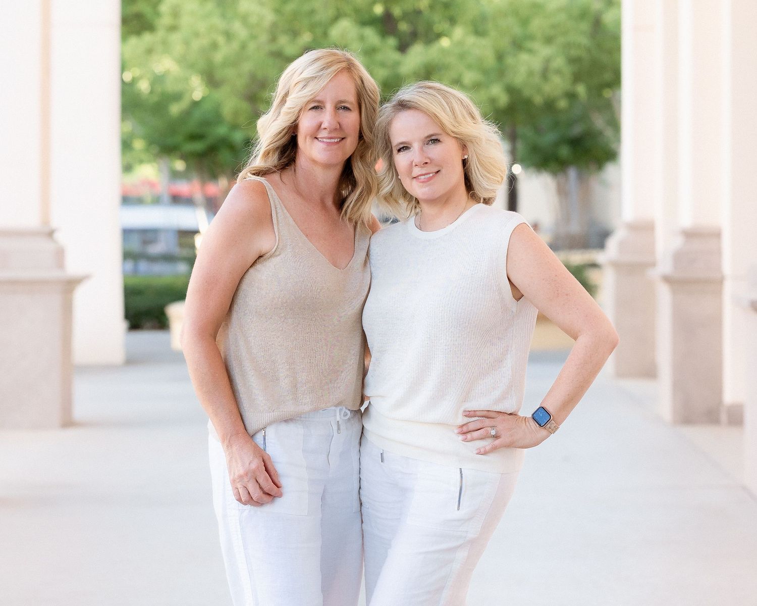 Two smiling women in white and beige outfits posing together outdoors near elegant columns.