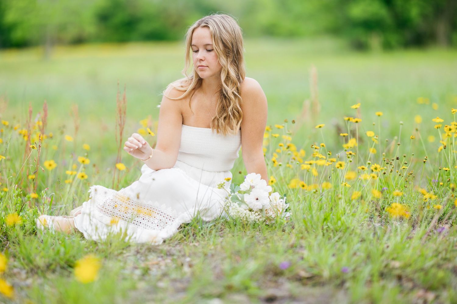 A person in a white dress sits gracefully in a summer meadow filled with yellow wildflowers and green grass.