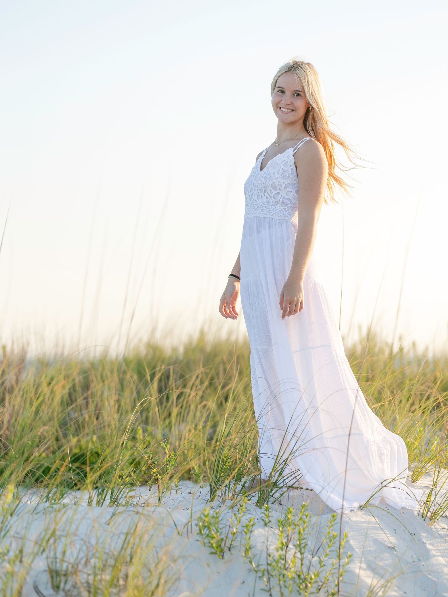 Ethereal white dress flows in the sea breeze as figure stands among coastal dunes at sunset.