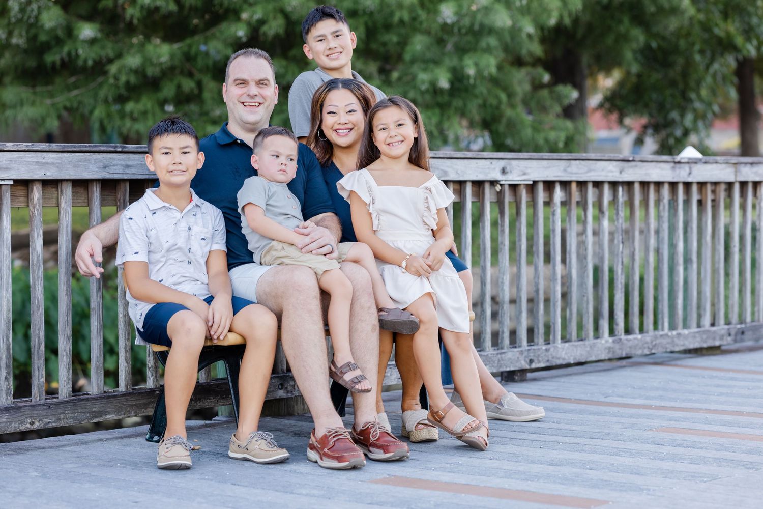 A family sits together on a wooden bridge railing during a summer outdoor photo session.