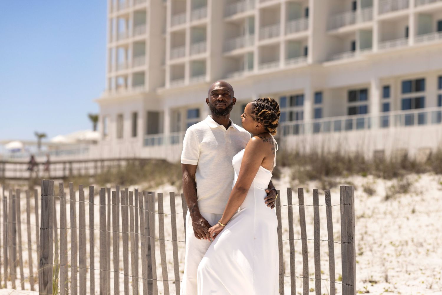 A couple in white attire holds hands on a sandy beach with a fence and resort building in the background.