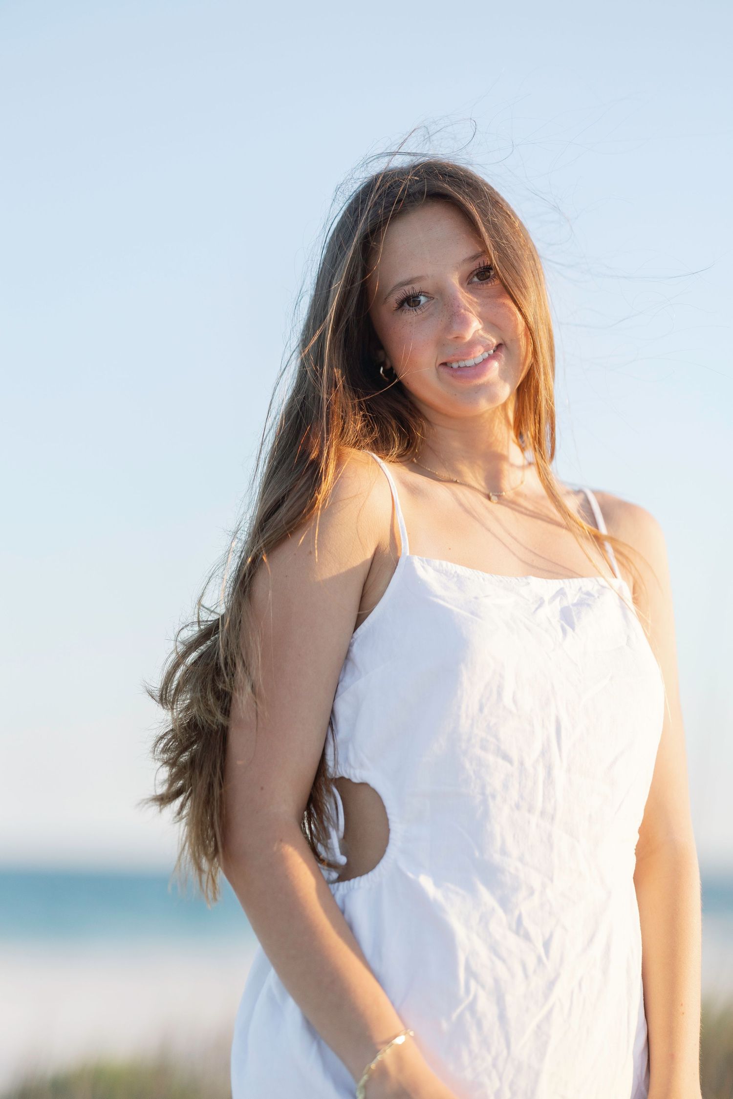 Woman in white dress poses playfully on sunny beach with wind-swept hair and natural lighting.