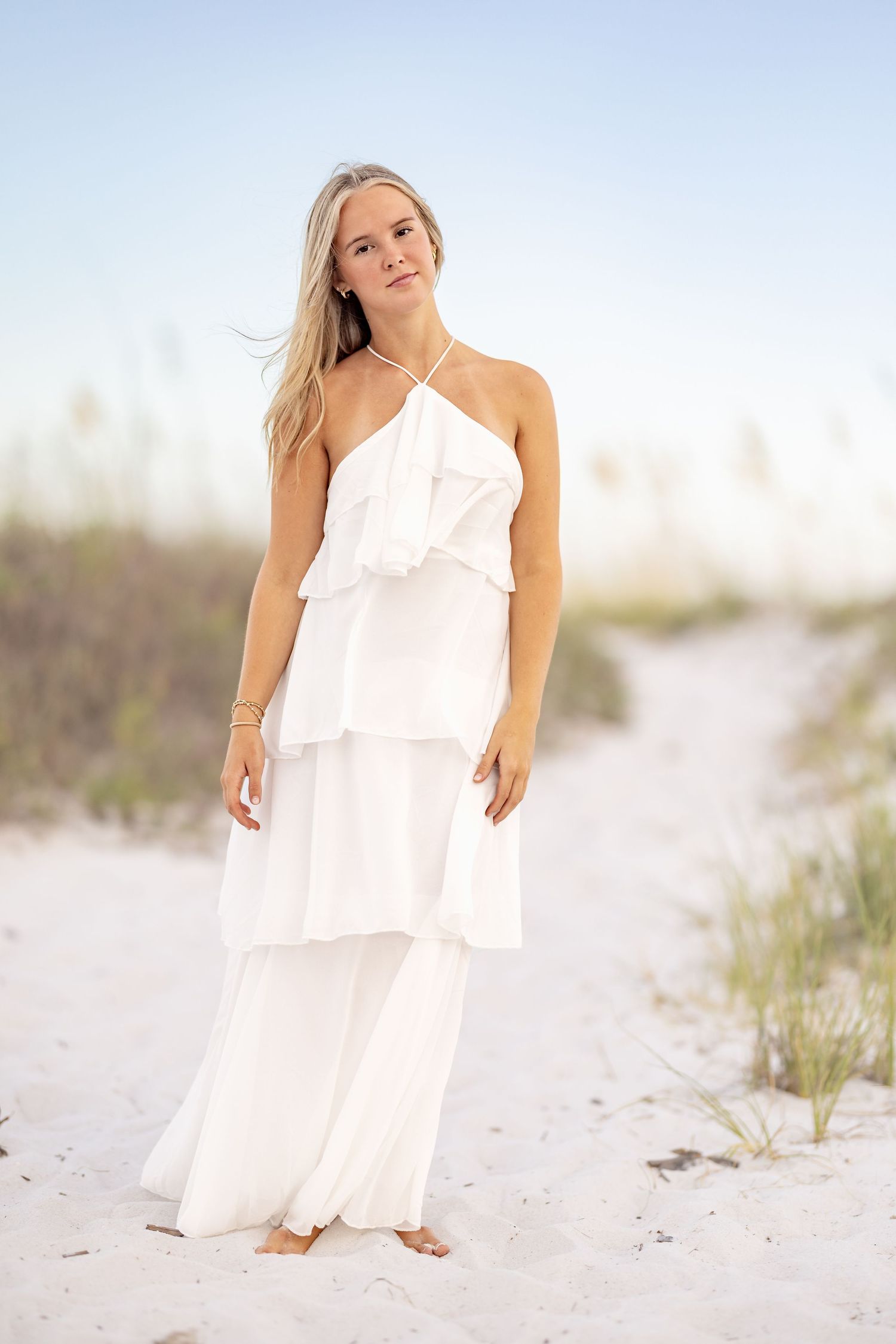 Young woman in white tiered ruffle halter maxi dress standing on sandy beach with dunes and clear sky.