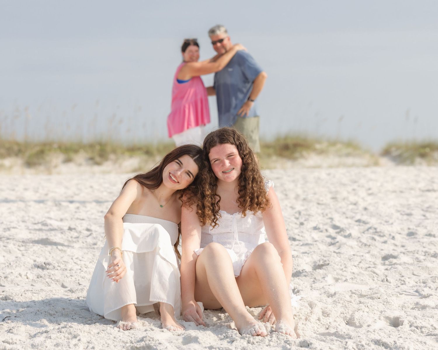 Two girls pose on white sand beach while parents stand behind them in a sunny family beach photo session.