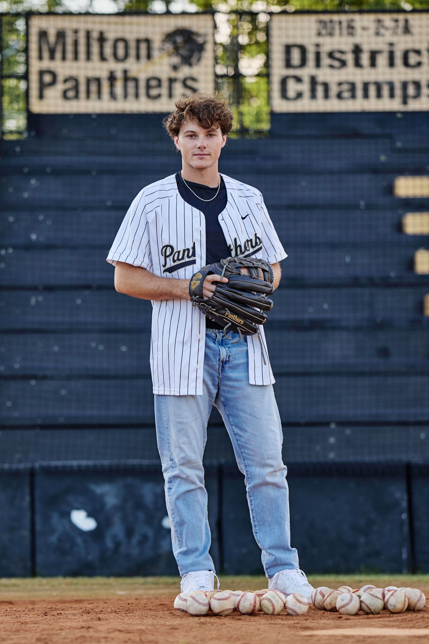 Young baseball player in Milton Panthers jersey holding a glove, standing on field near District Champions sign.