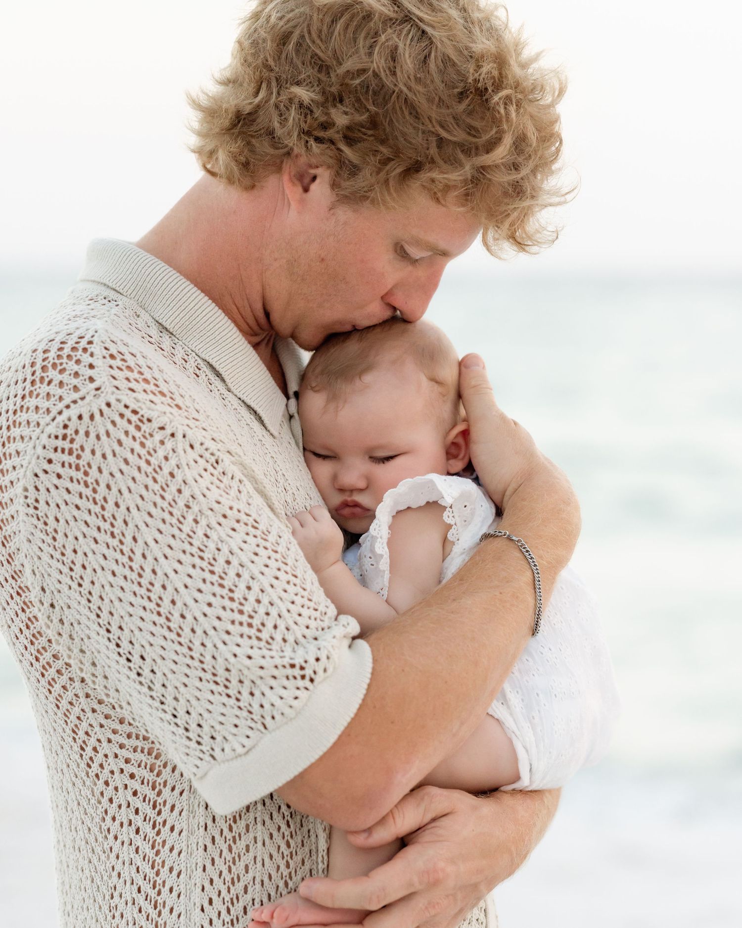 Tender moment of curly-haired father kissing sleeping newborn baby on beach.
