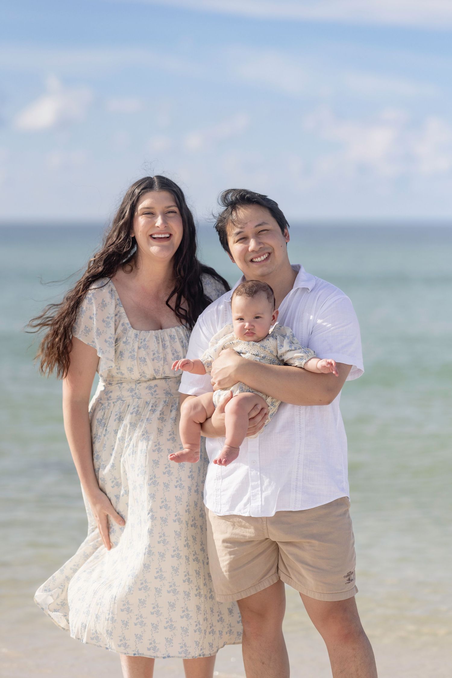 Happy family of three posing on a sunny beach, parents smiling with their baby, ocean waves in the background.
