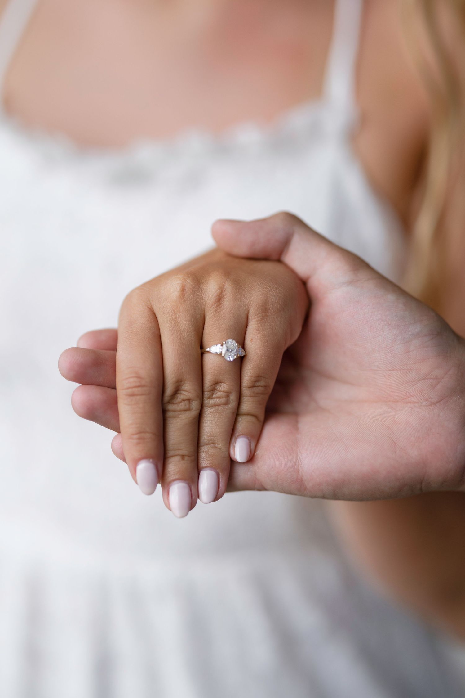 Close-up of a diamond engagement ring on a woman's hand with pink nails, held gently by another person.