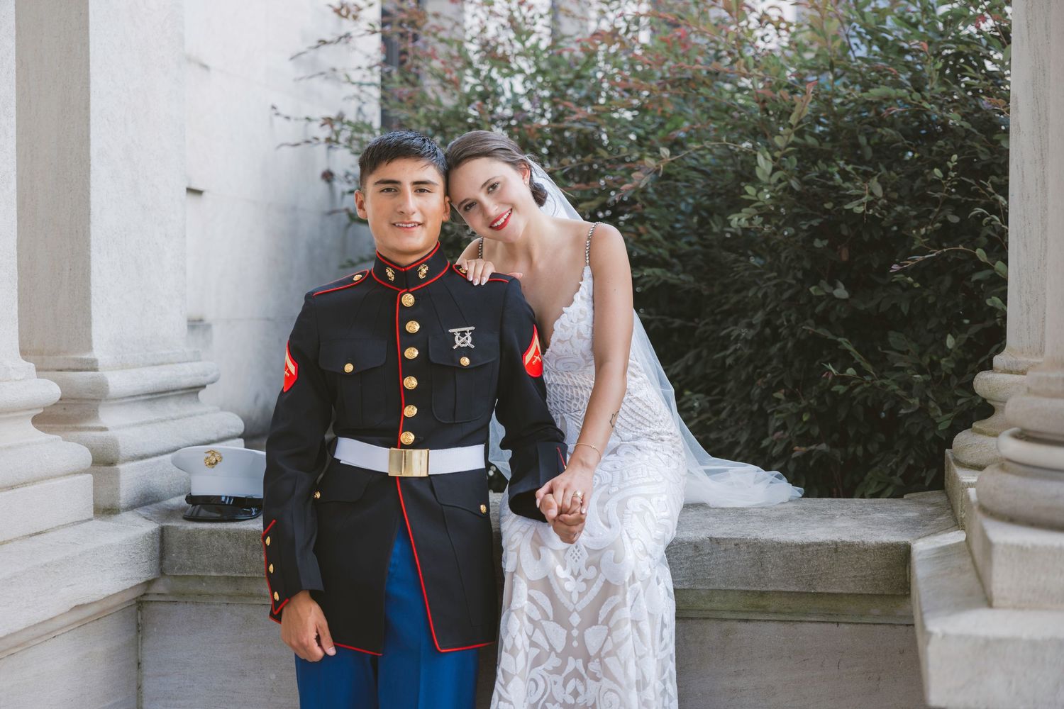 A Marine in dress blues stands with a bride in a white lace gown at an elegant outdoor wedding venue.