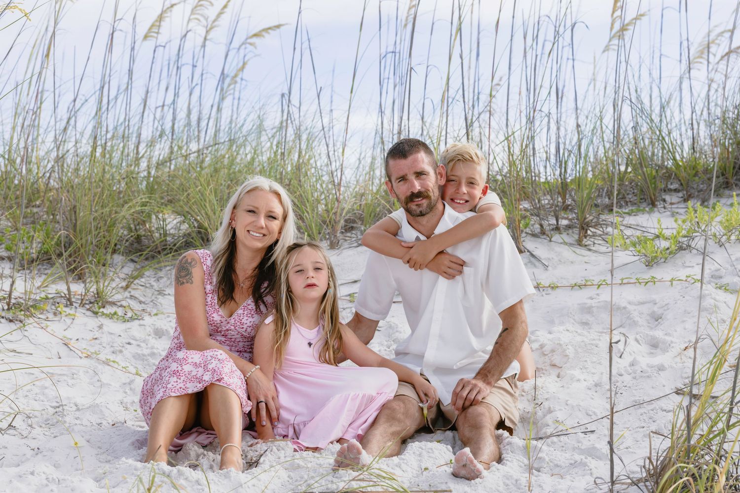 Family of four smiling together on sandy beach dunes with sea grass, wearing casual summer outfits on a sunny day.