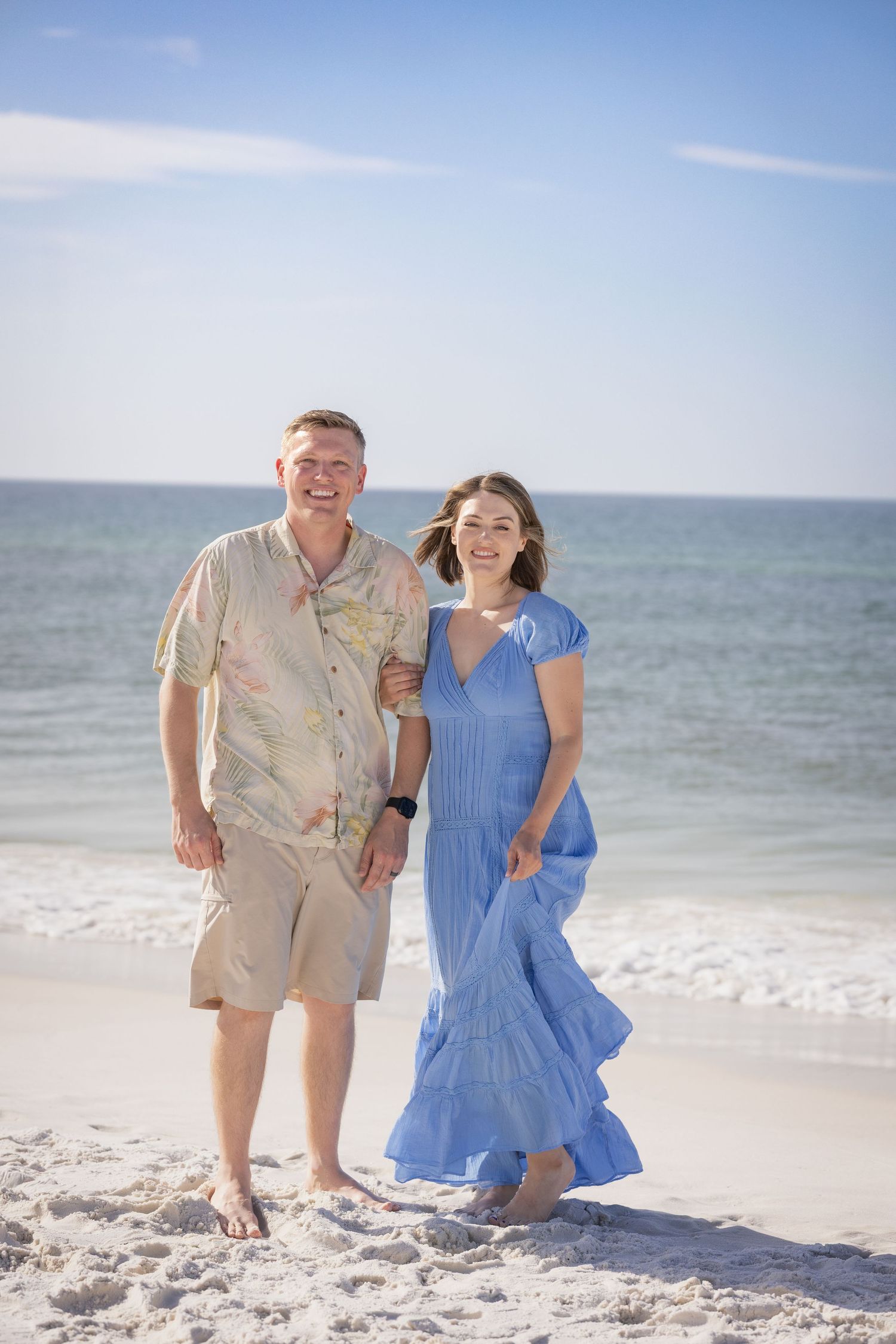 A smiling couple walks barefoot on a sunny white sand beach, woman in blue maxi dress, man in Hawaiian shirt.