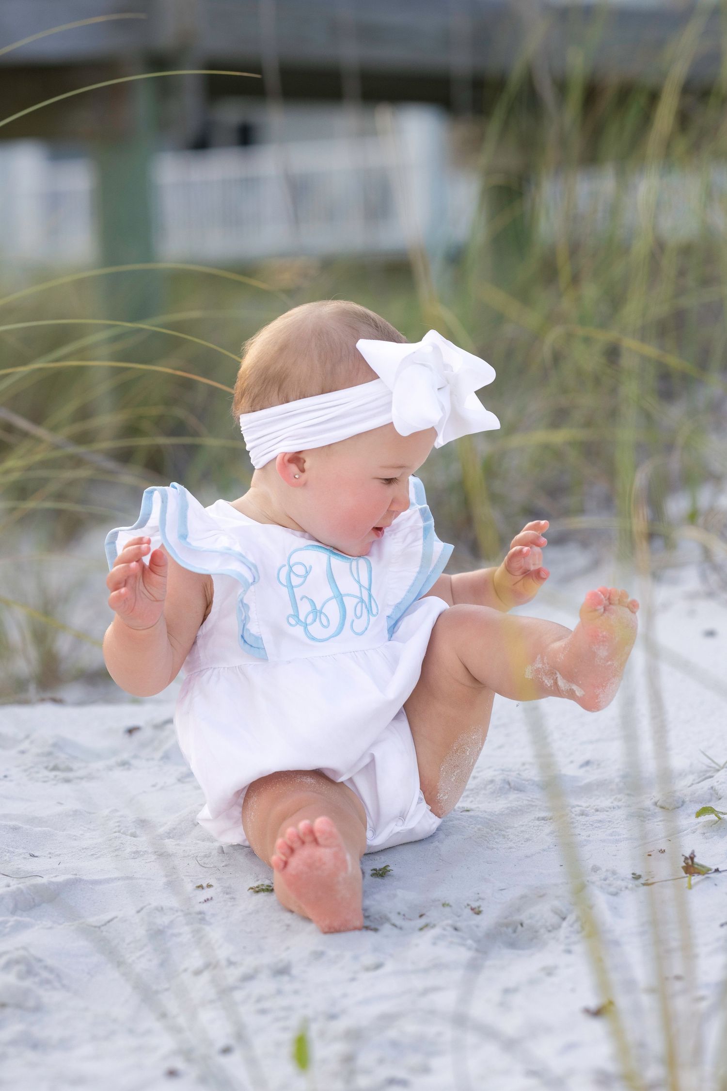Baby girl in white monogrammed romper and bow headband sitting on white sand beach dunes.