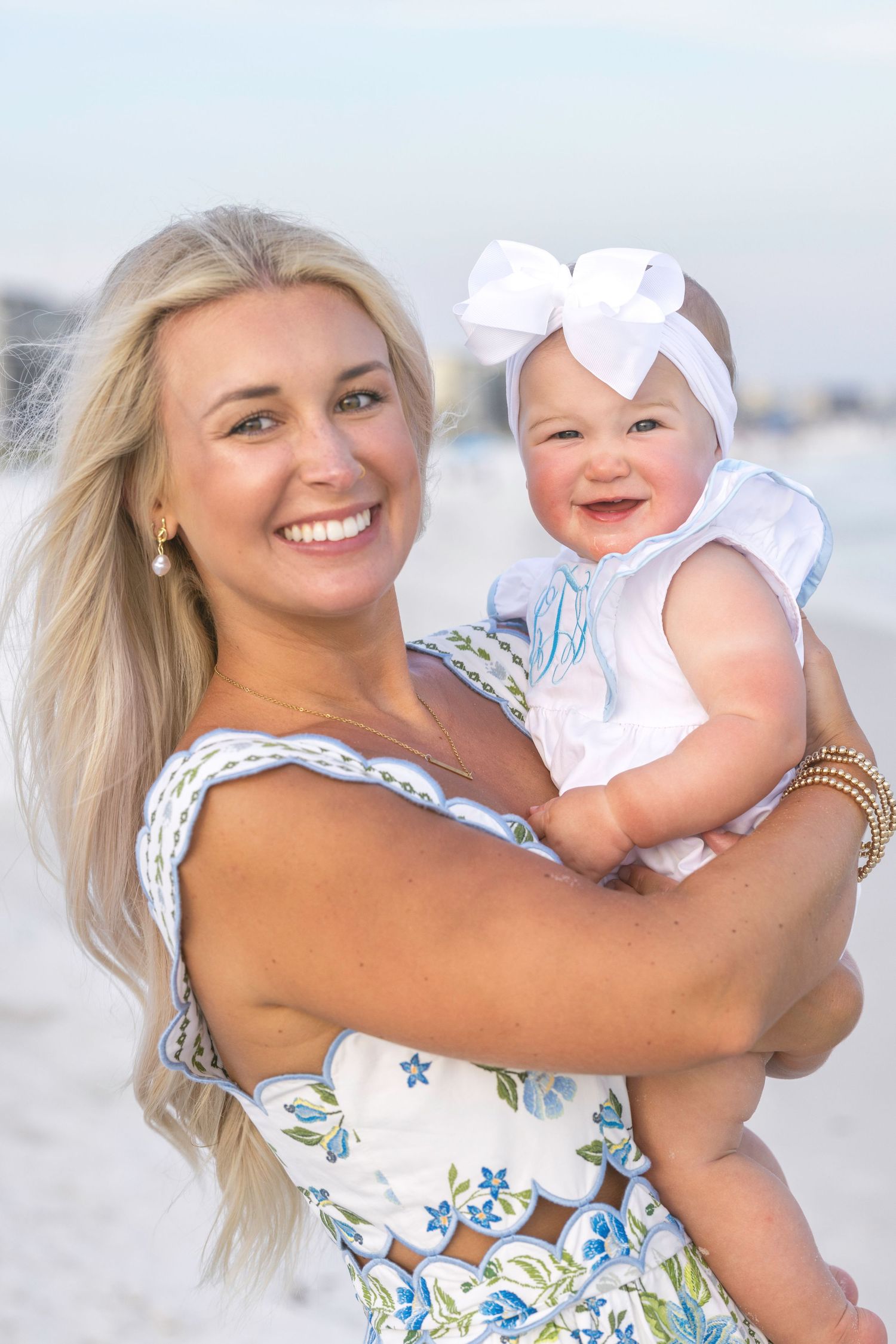 Smiling blonde woman holding a happy baby girl in a white bow headband on a sunny beach.