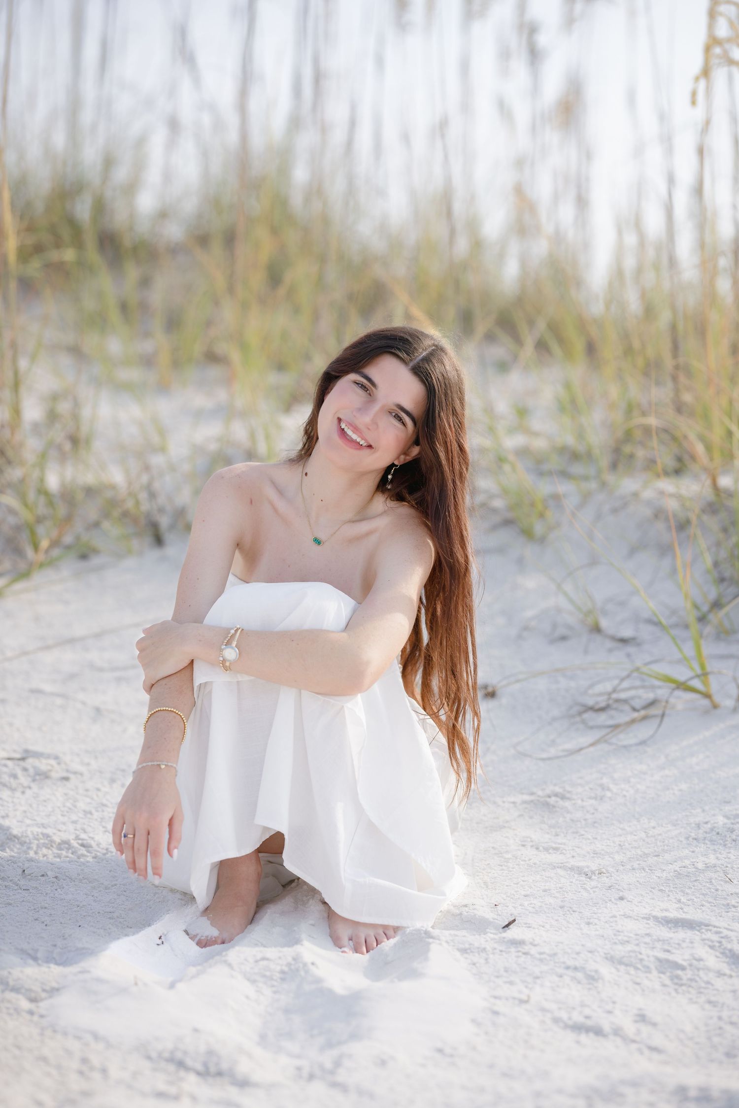 Smiling young woman in white strapless dress sitting on sandy beach dunes with sea grass.