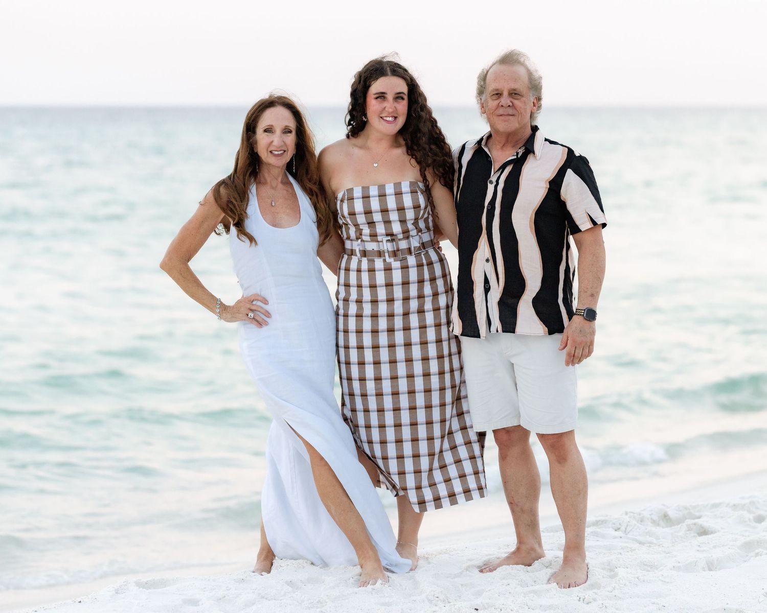 Three people smiling together on a sunny beach with turquoise water, wearing summer outfits for a family beach photo.