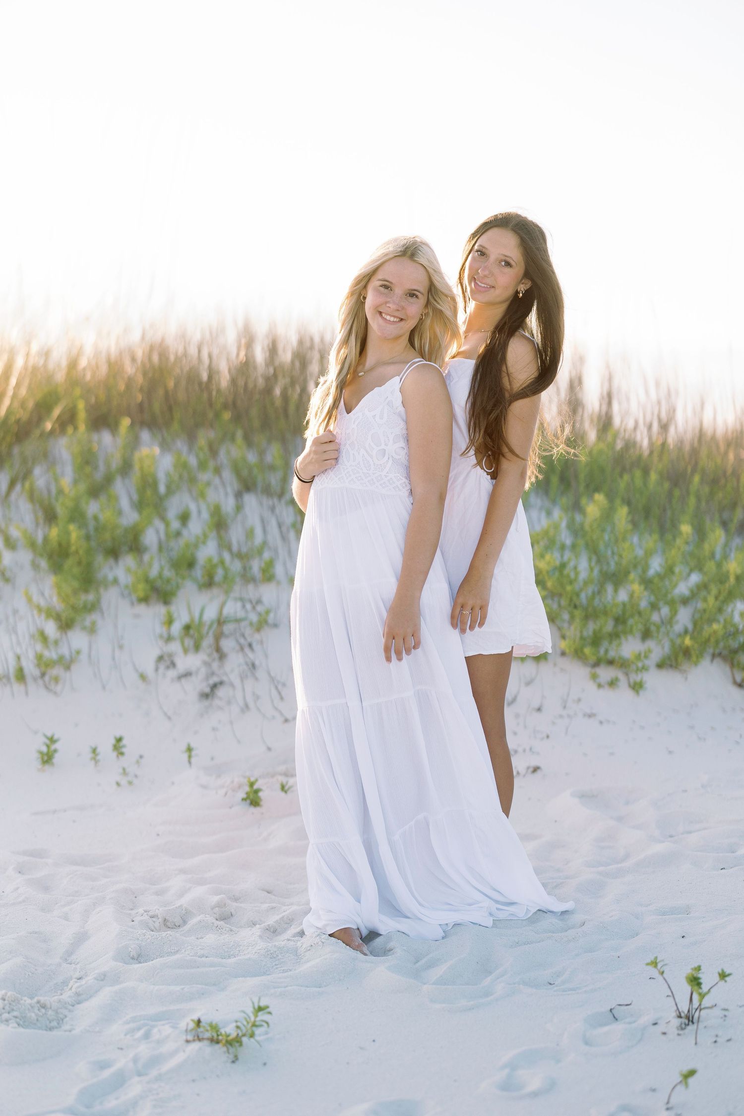 Two friends in white dresses pose together on sandy beach with dune grass.