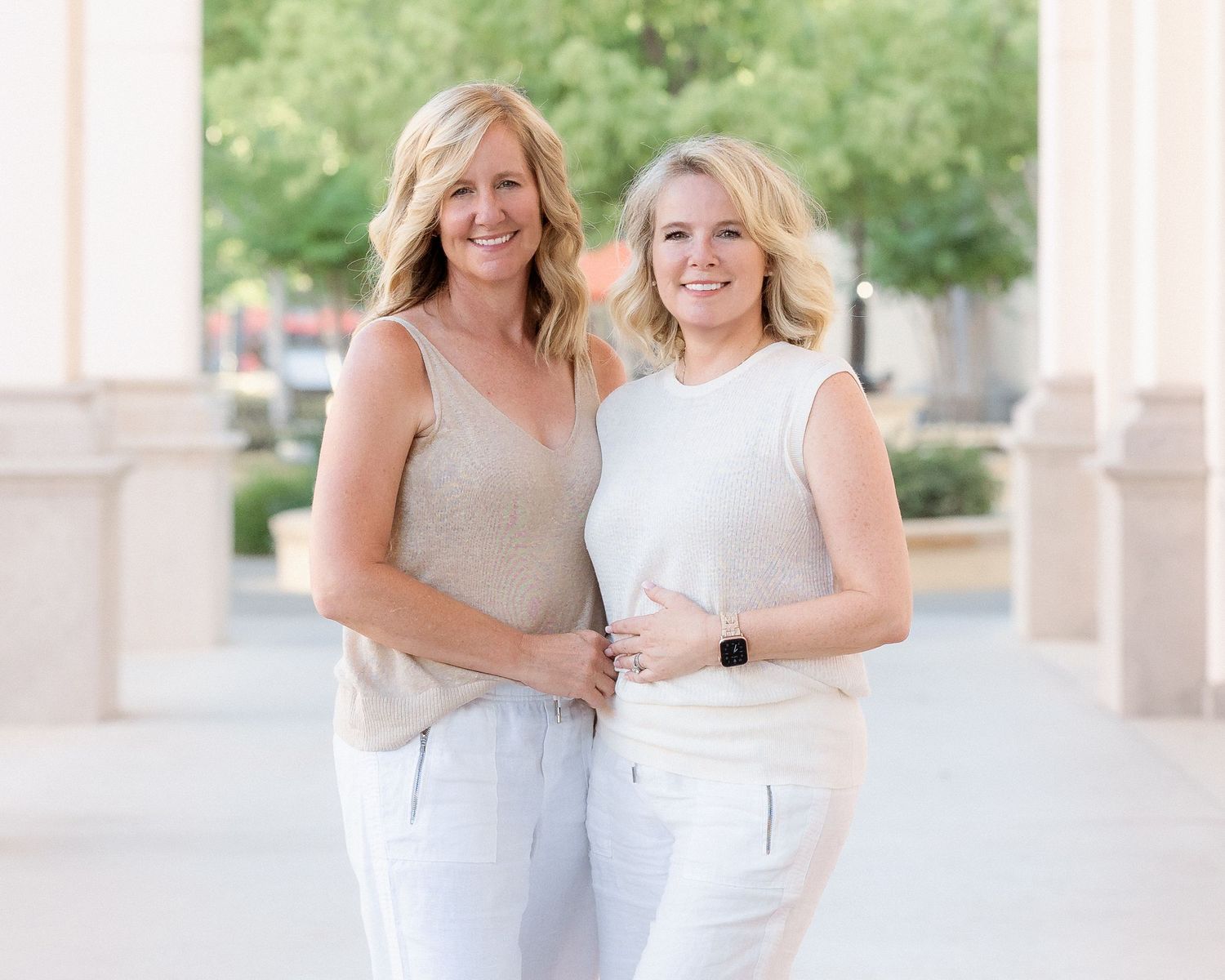 Two smiling blonde women in light outfits pose together outdoors near columns.