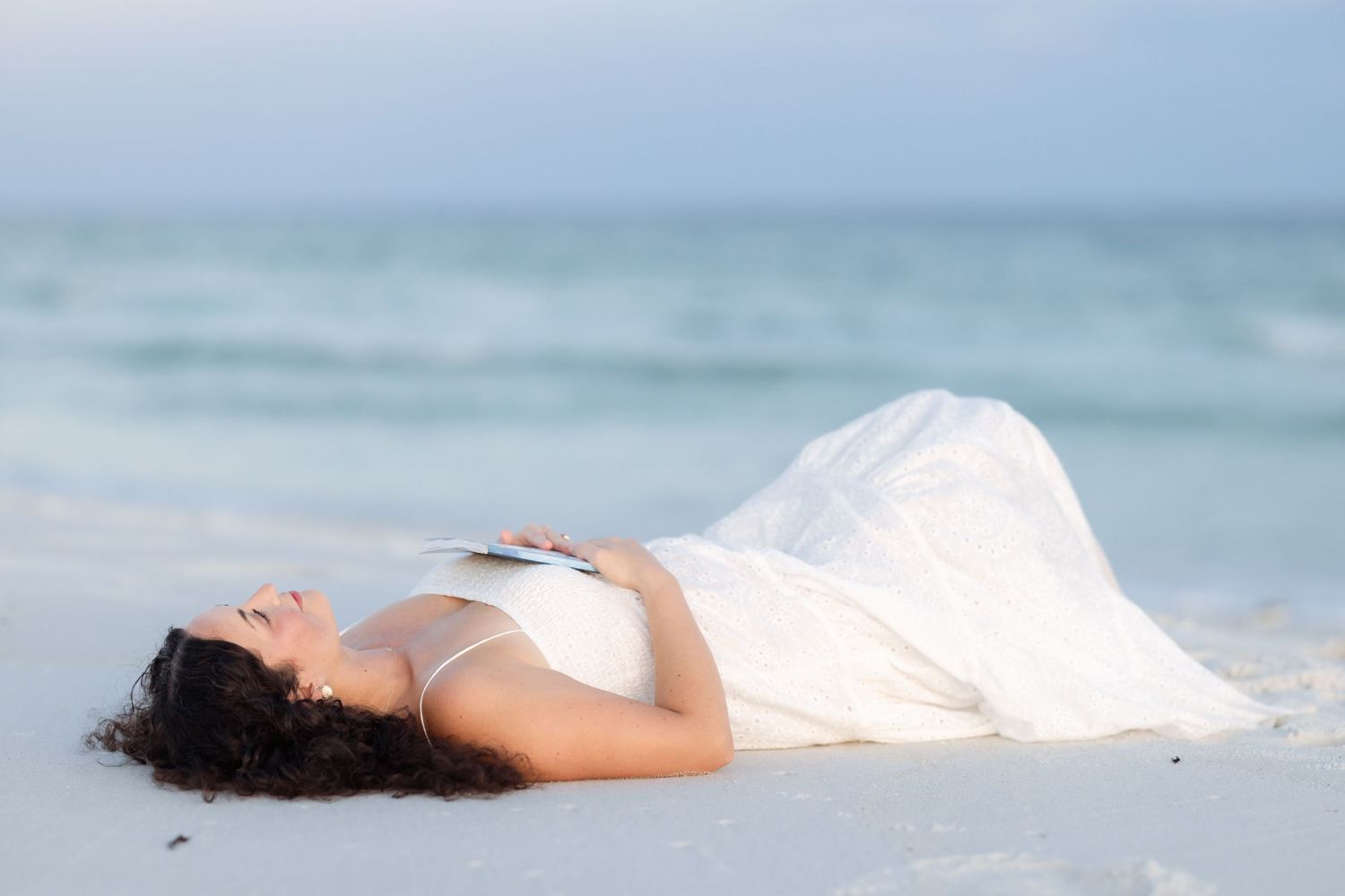 Pregnant woman in white dress lying on white sand beach with ocean waves in background.