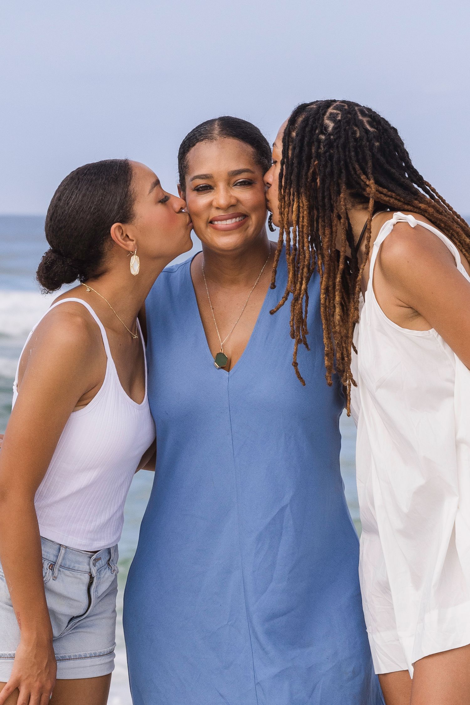 Three women at the beach, two kissing a smiling woman in a blue dress on her cheeks in a joyful outdoor moment.