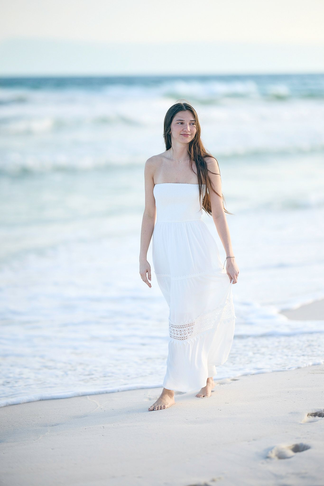 A young woman in a white strapless maxi dress walks barefoot along a sandy beach with ocean waves behind her.