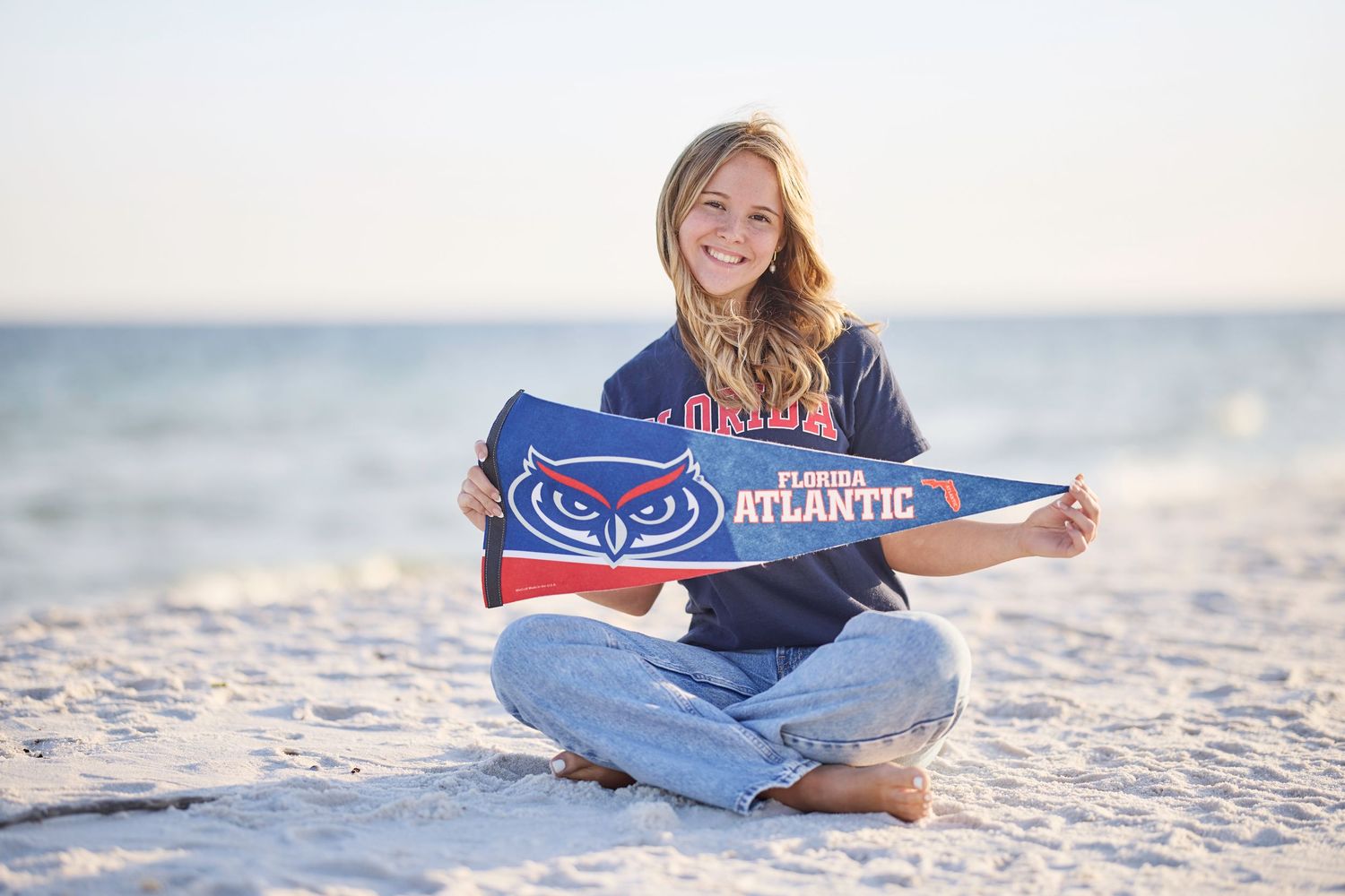 Smiling girl holding Florida Atlantic University pennant flag on sandy beach in FAU hoodie.