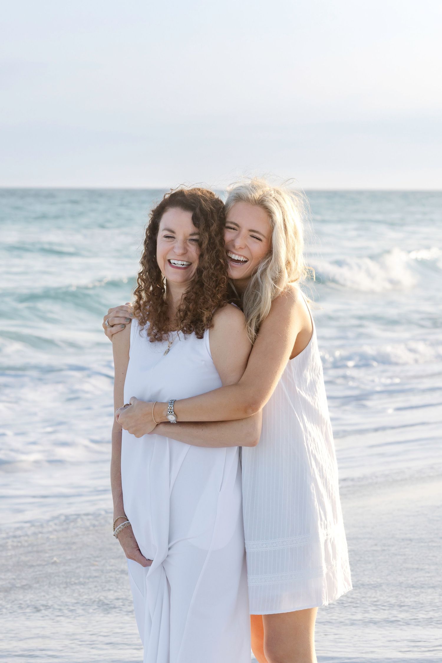 Two women in white dresses laughing and embracing on a sunny beach with ocean waves in the background.