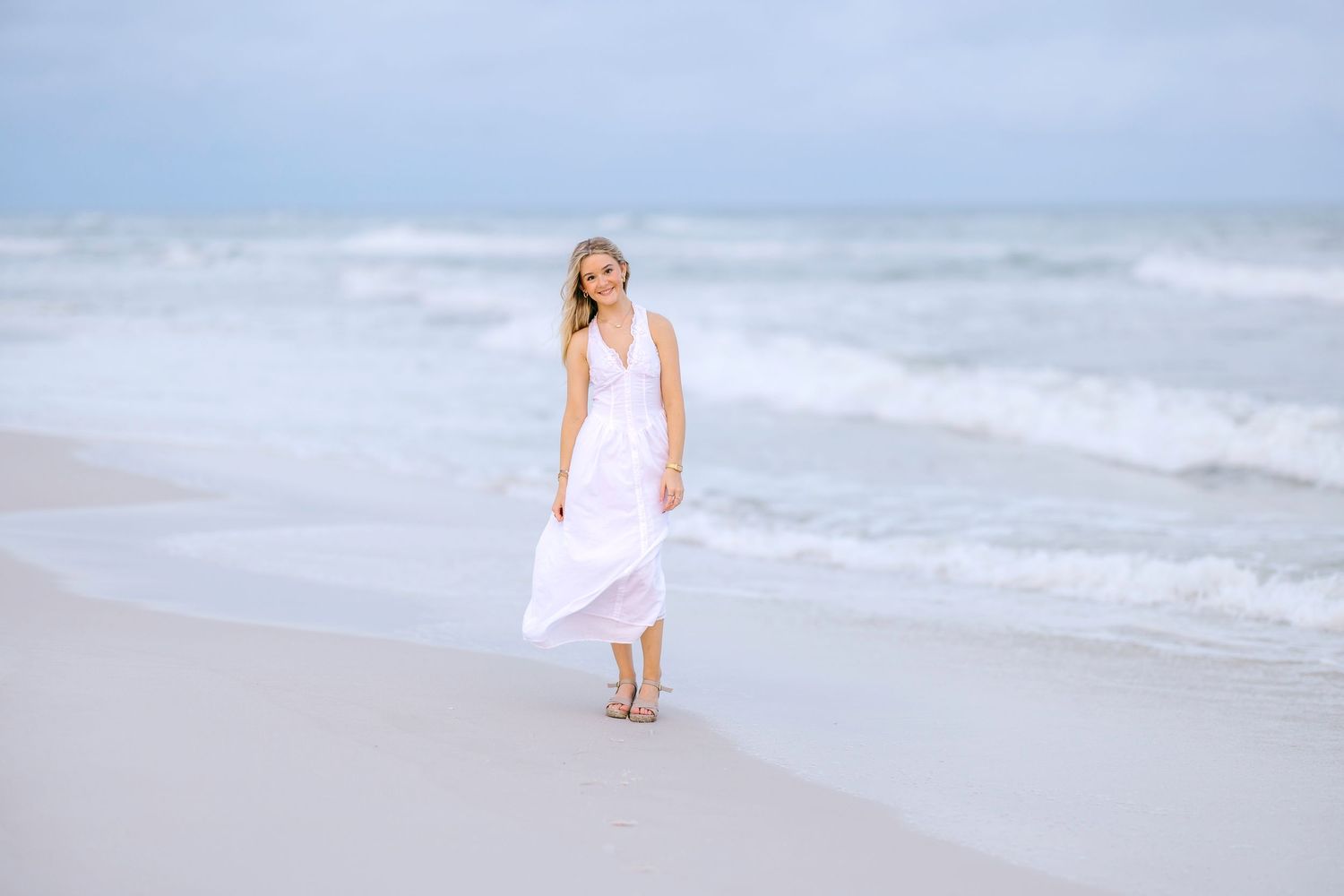 Woman in white flowing dress walking on sandy beach with ocean waves, smiling outdoors.