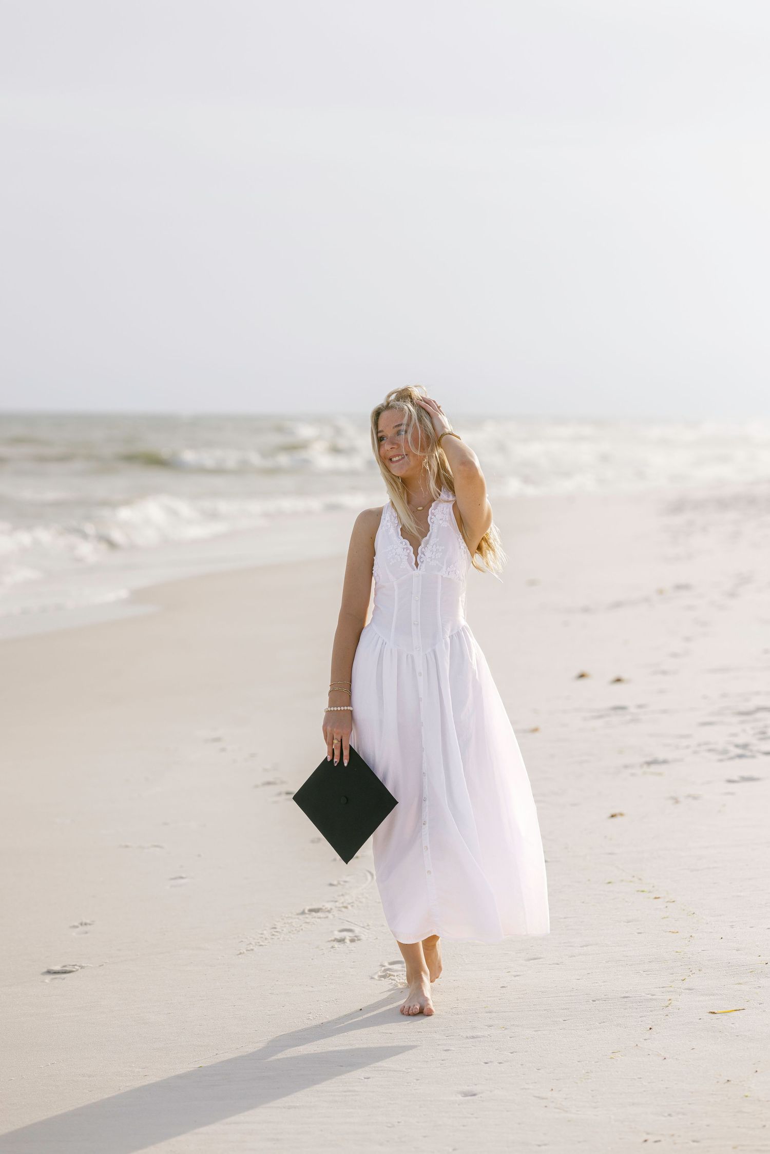 Young woman in white maxi dress holding graduation cap, walking barefoot on a sandy beach with ocean waves.