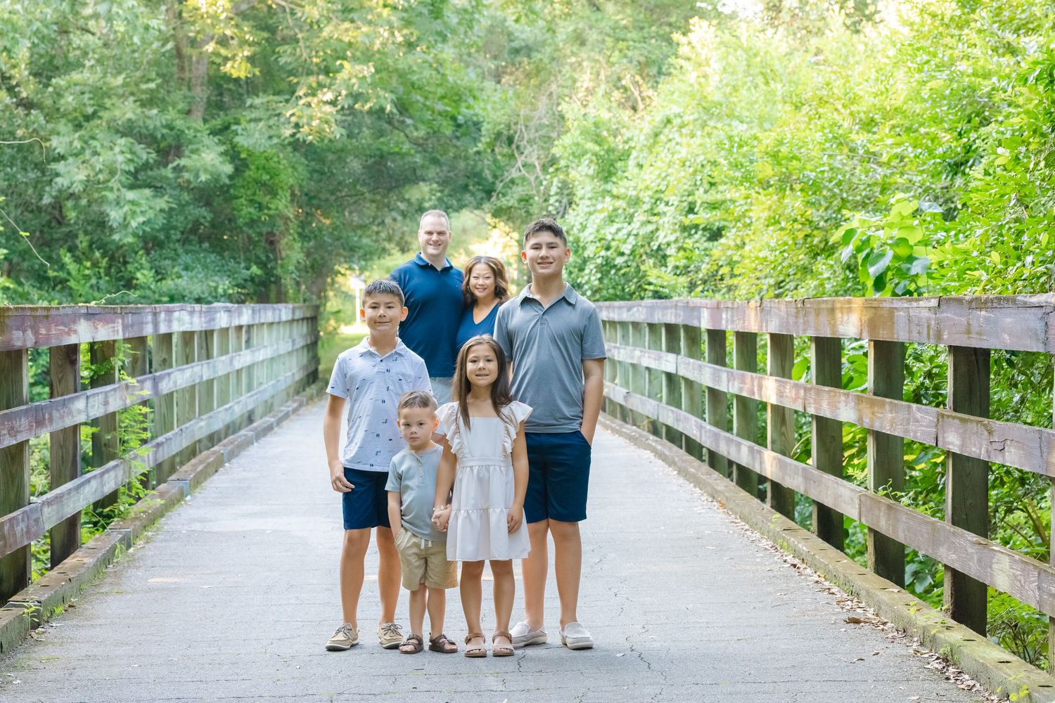 Family of six posing on a wooden bridge surrounded by lush green trees for an outdoor portrait.