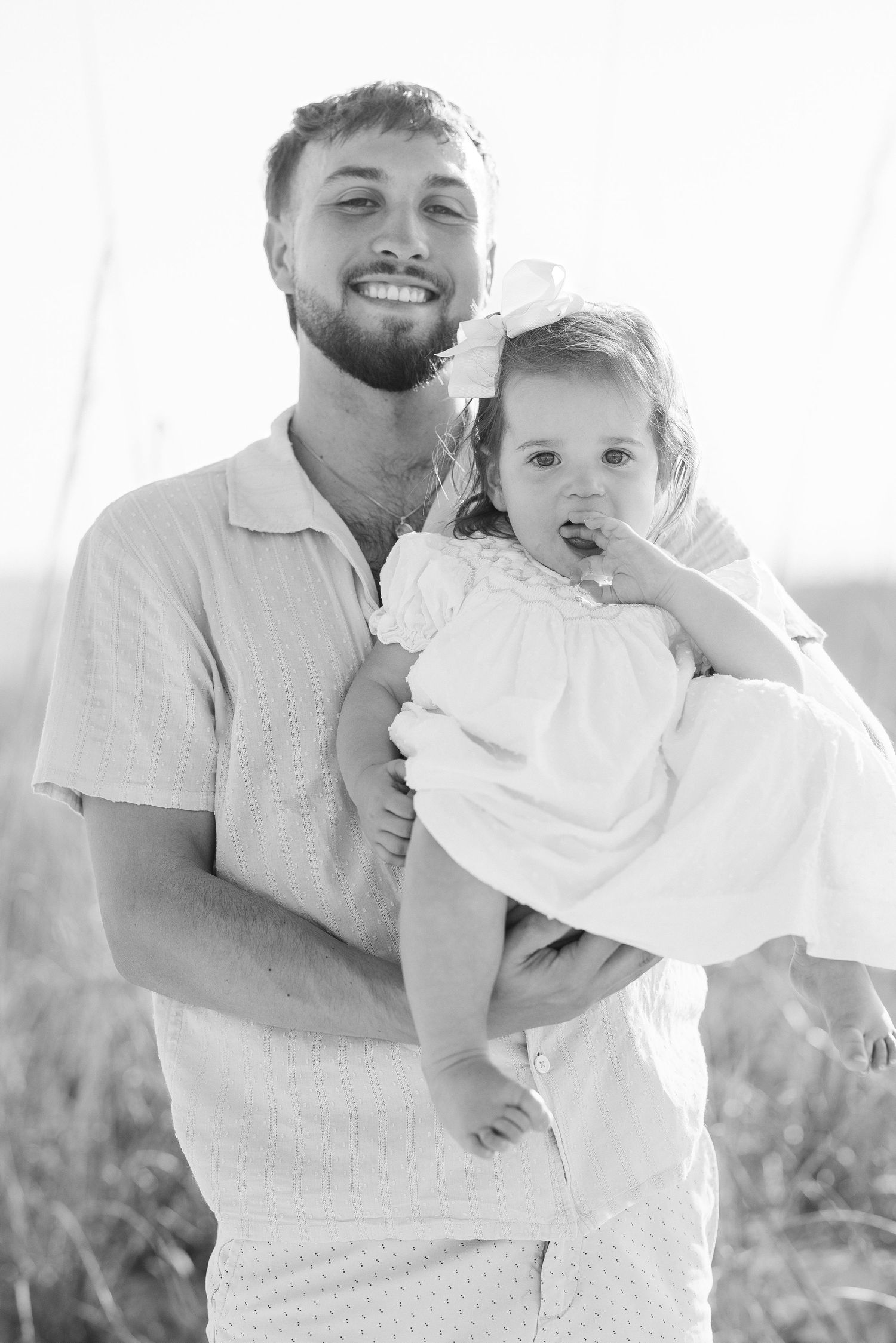 Black and white photo of a smiling bearded man holding a baby girl with a bow in her hair outdoors.
