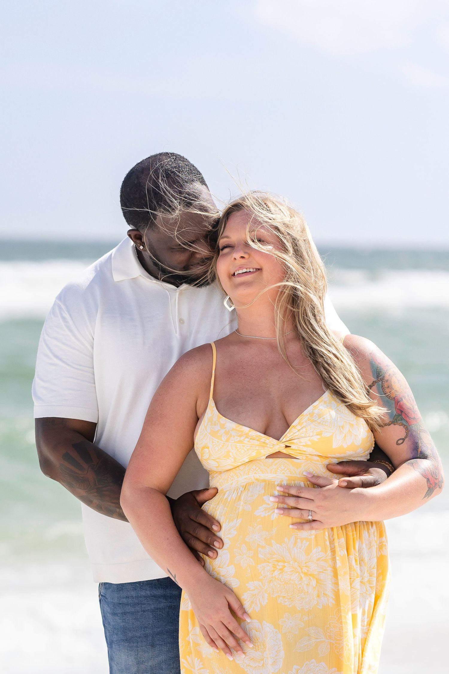 Interracial couple sharing a tender beach moment, pregnant woman in yellow floral dress smiling as partner embraces her.