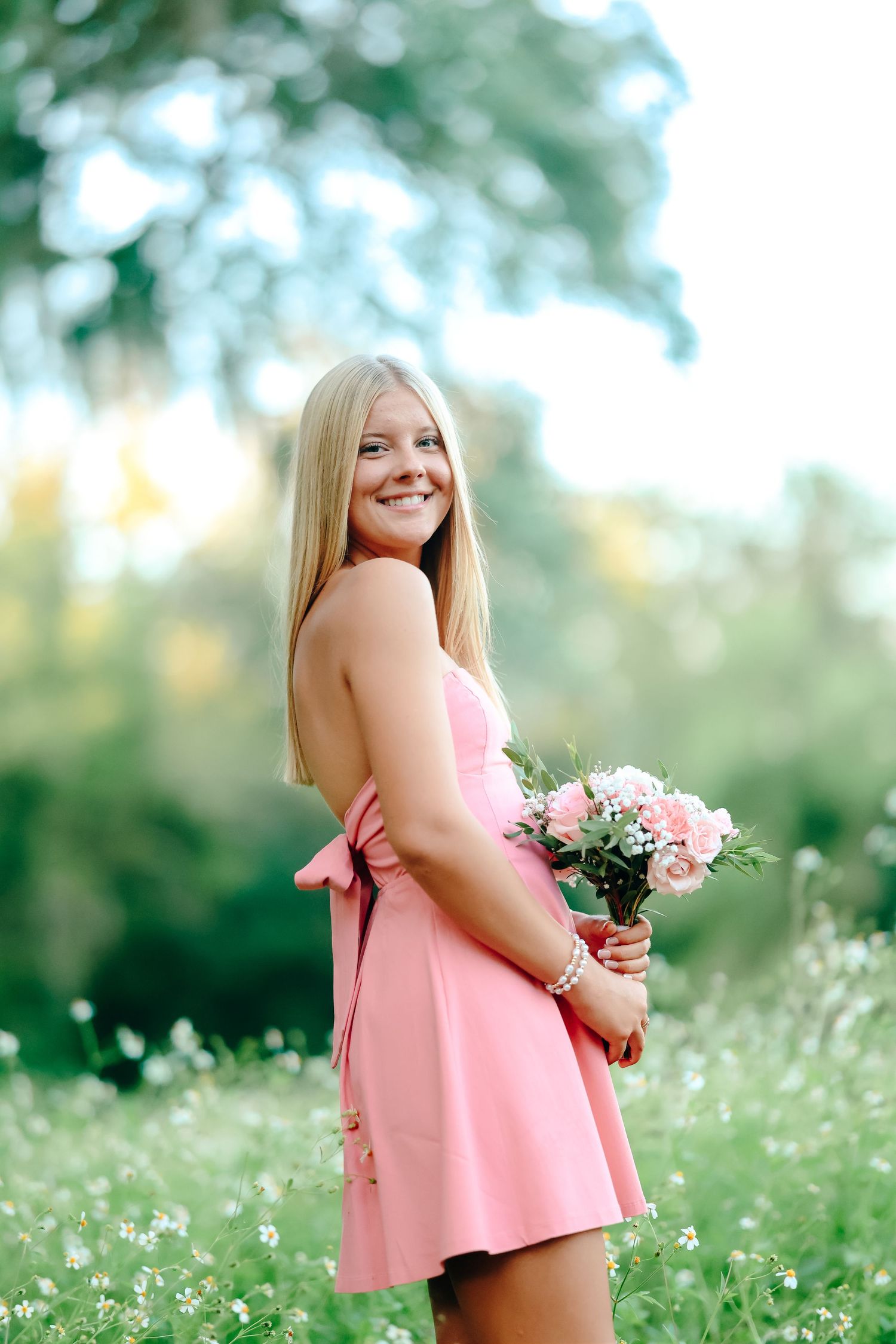 Smiling blonde woman in pink dress holding bouquet of pink roses in a lush green outdoor setting.