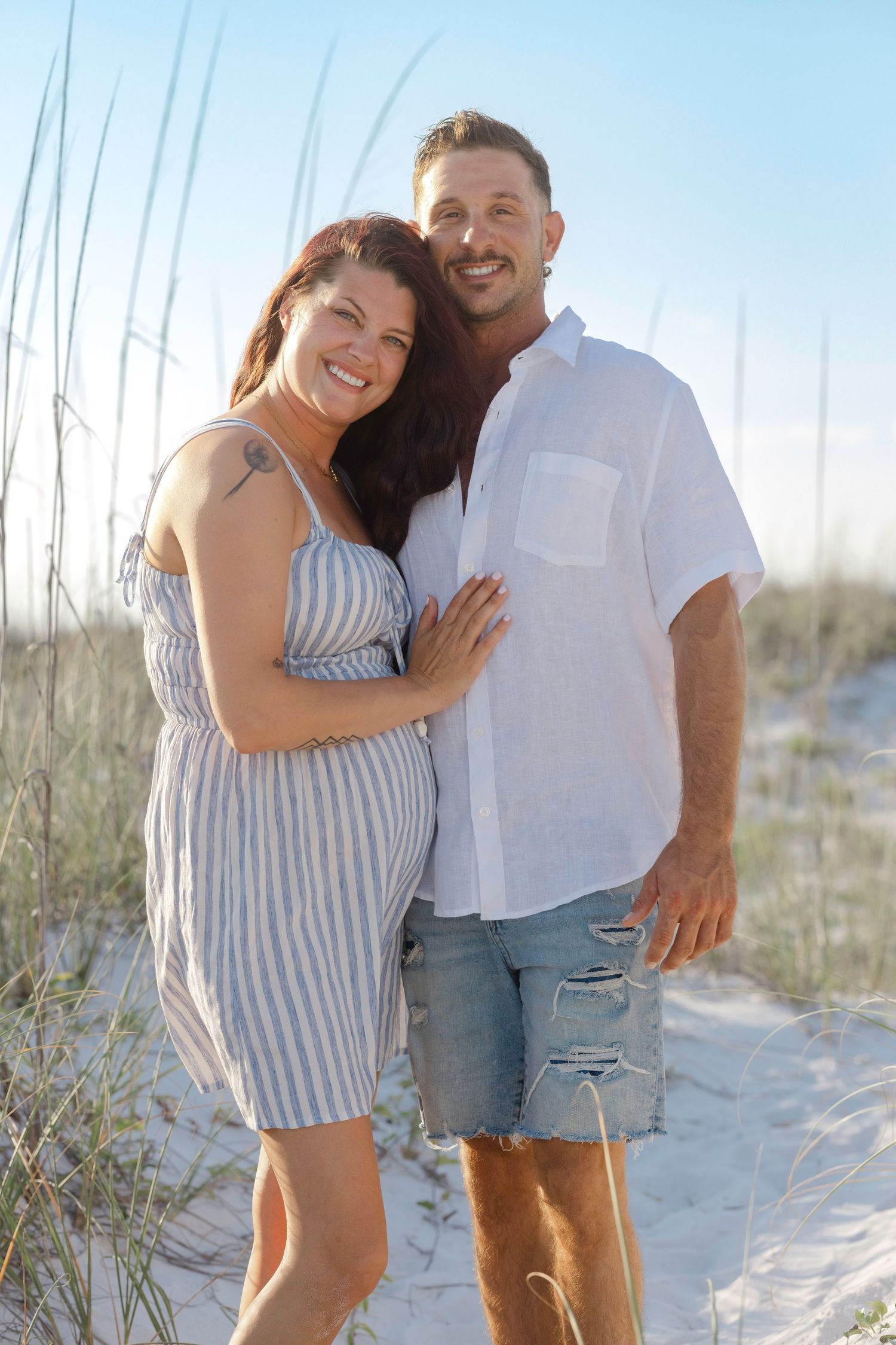 Smiling couple posing on sandy beach dunes, woman in striped dress and man in white shirt and denim shorts.