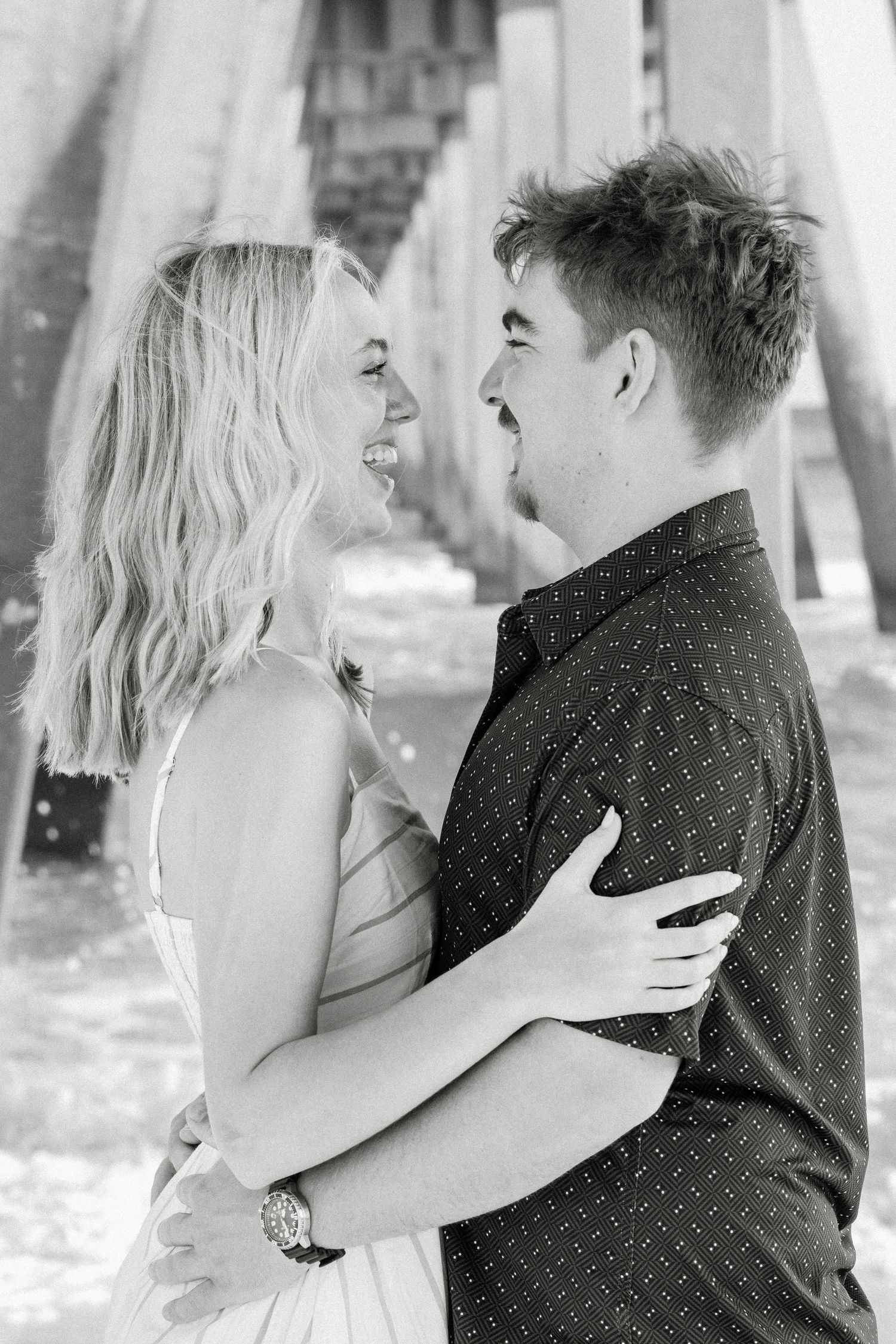 Black and white photo of a happy young couple embracing and laughing together under a beach pier.
