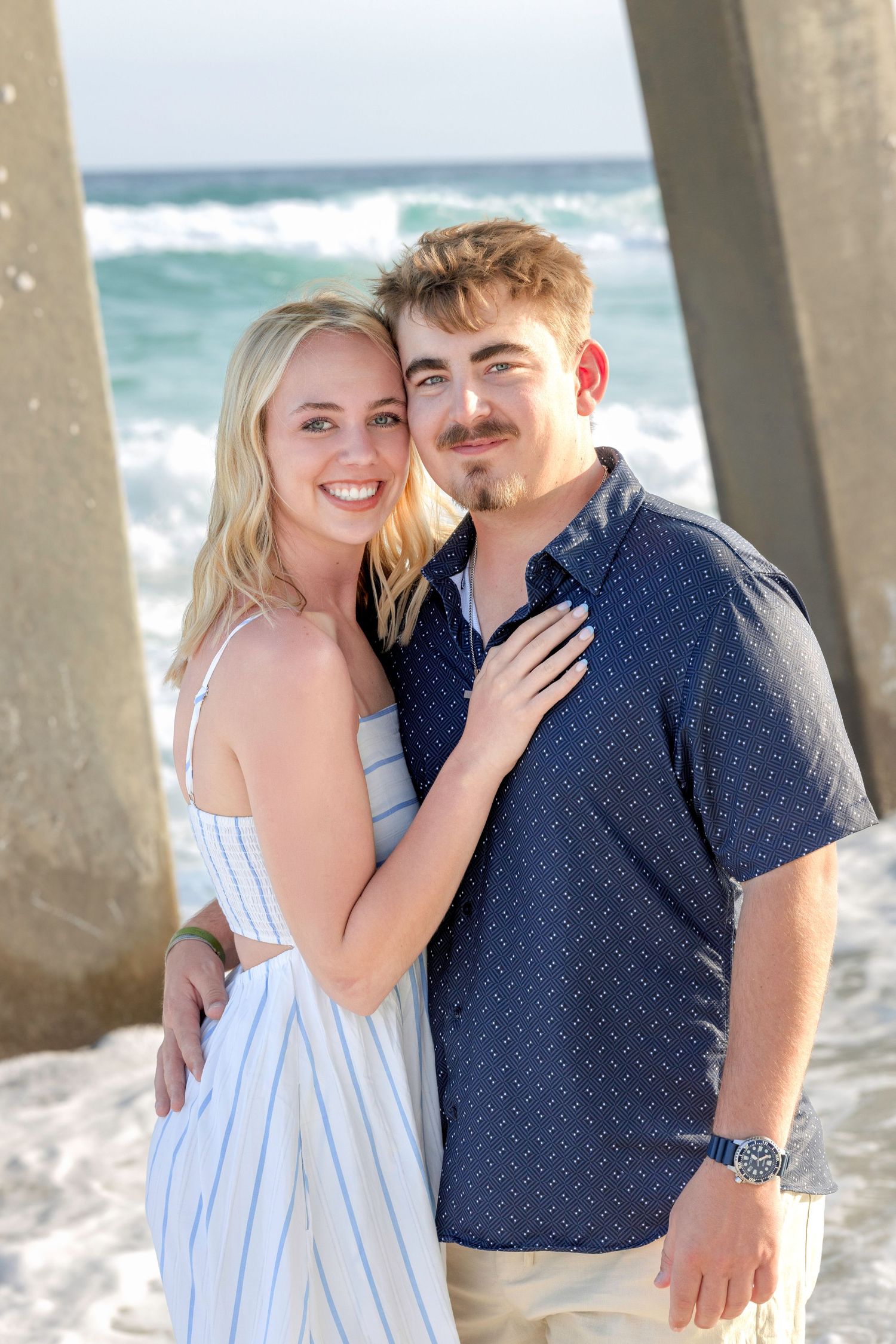 Happy couple embracing on beach under pier with ocean waves, woman in white dress and man in blue shirt.
