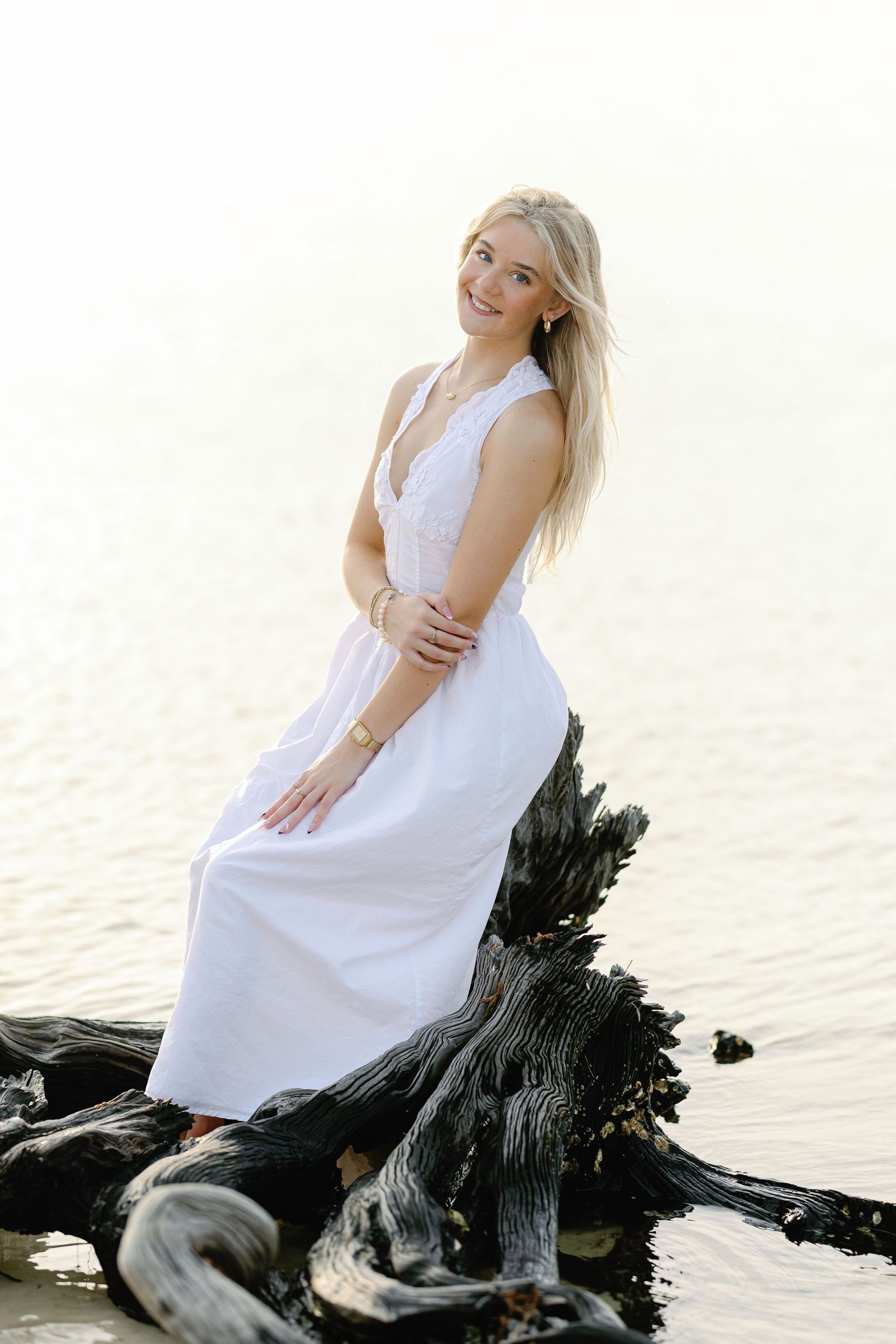 White maxi dress contrasts dramatically against dark driftwood on a serene beach.