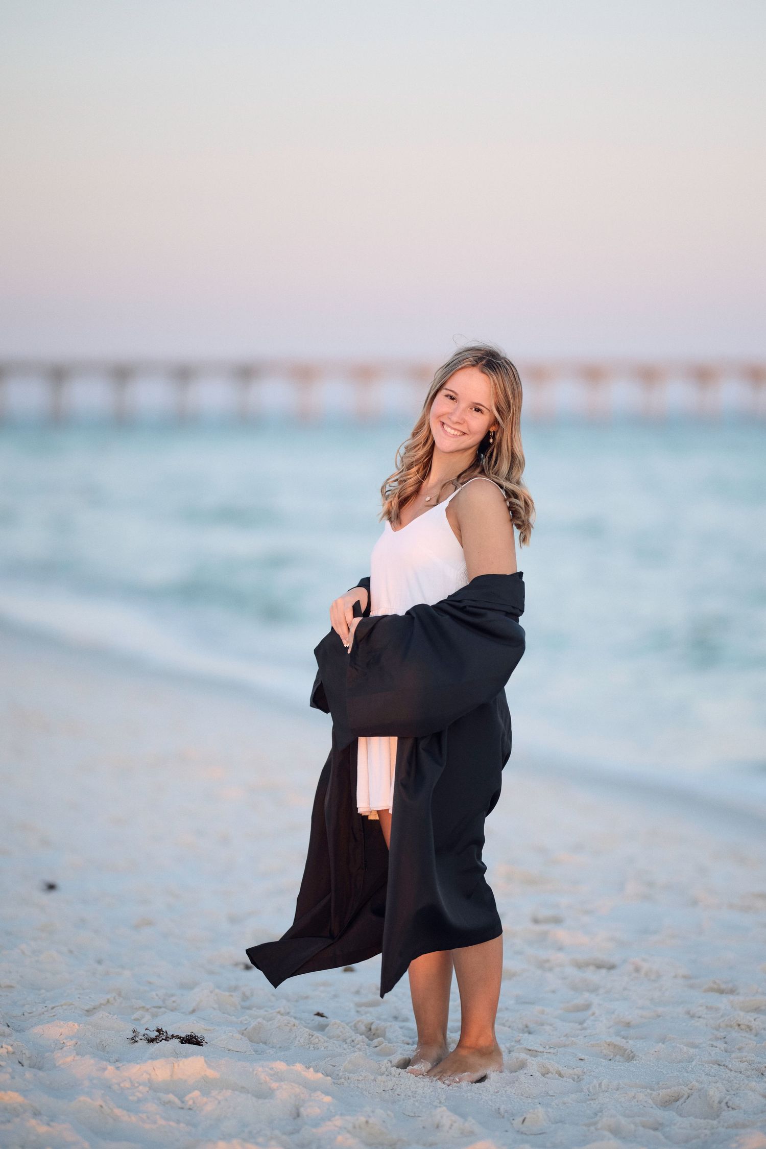 A person in a white dress and black cardigan poses on a beach at sunset with a pier in the background.