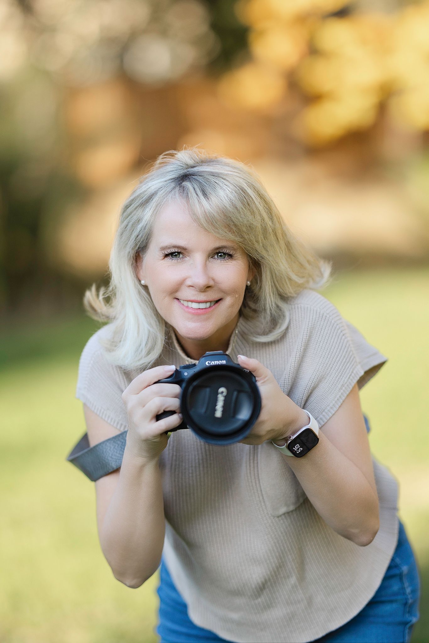 Professional photographer in beige sweater and jeans holds camera while smiling in outdoor setting with bokeh background.
