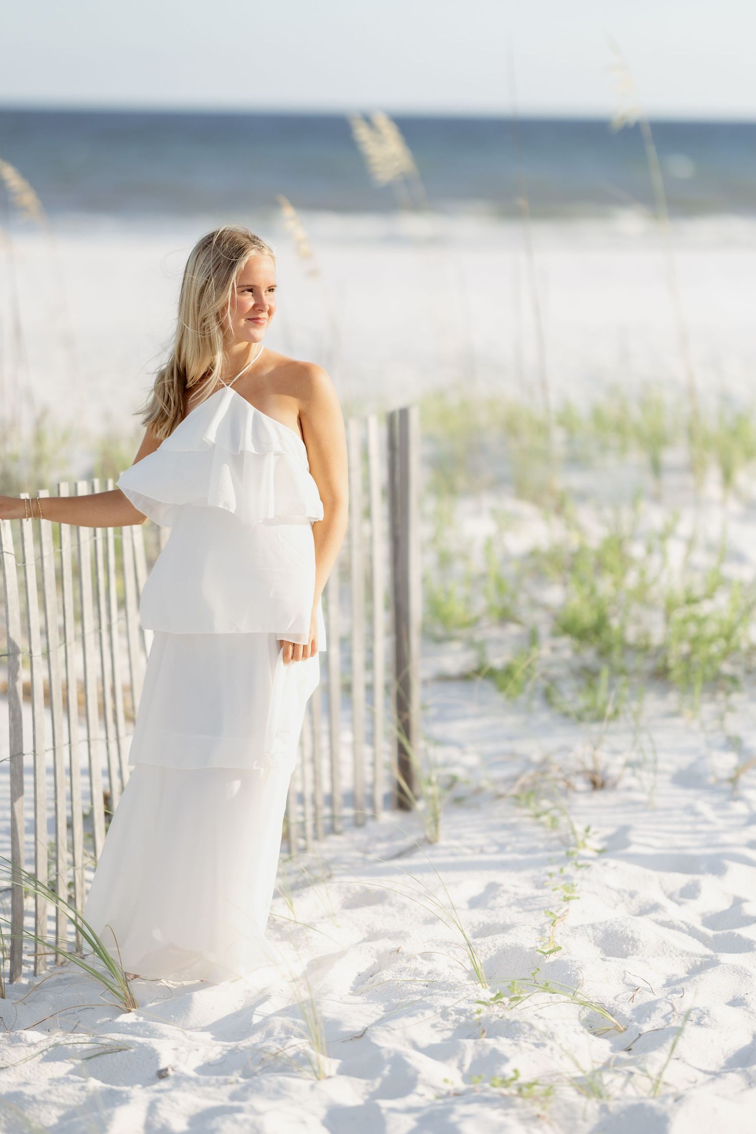 A blonde woman in a white halter maxi dress poses by a beach fence on white sand dunes with sea oats.