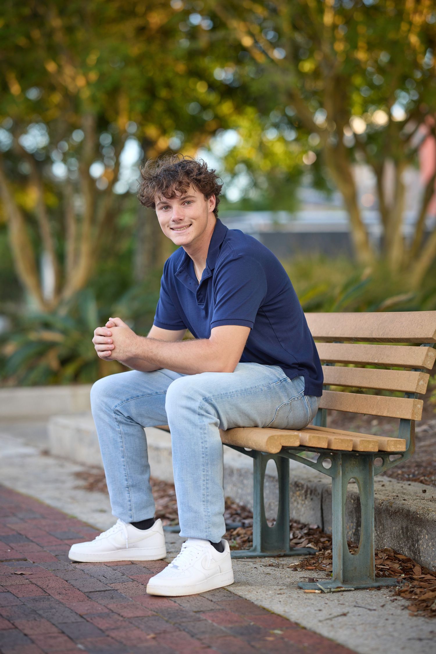 Young man in navy polo and jeans smiling on a wooden bench outdoors, surrounded by lush green trees.