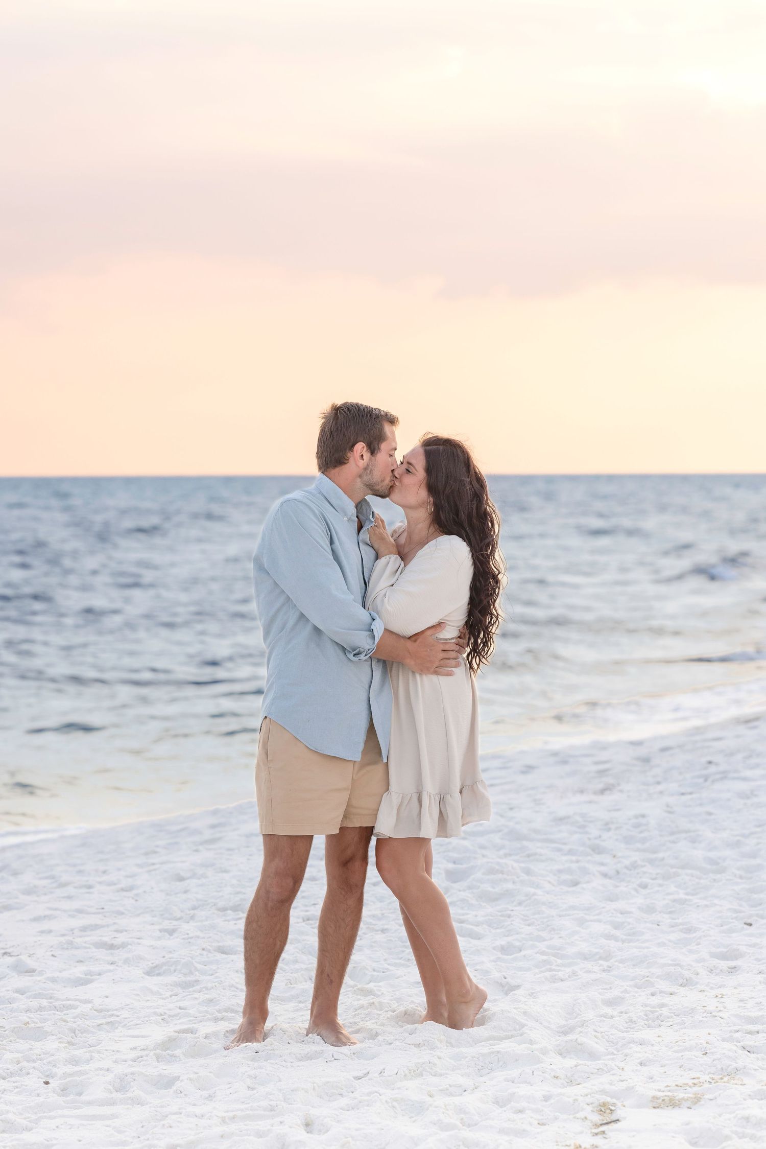 A couple shares a romantic kiss on a white sandy beach at sunset.