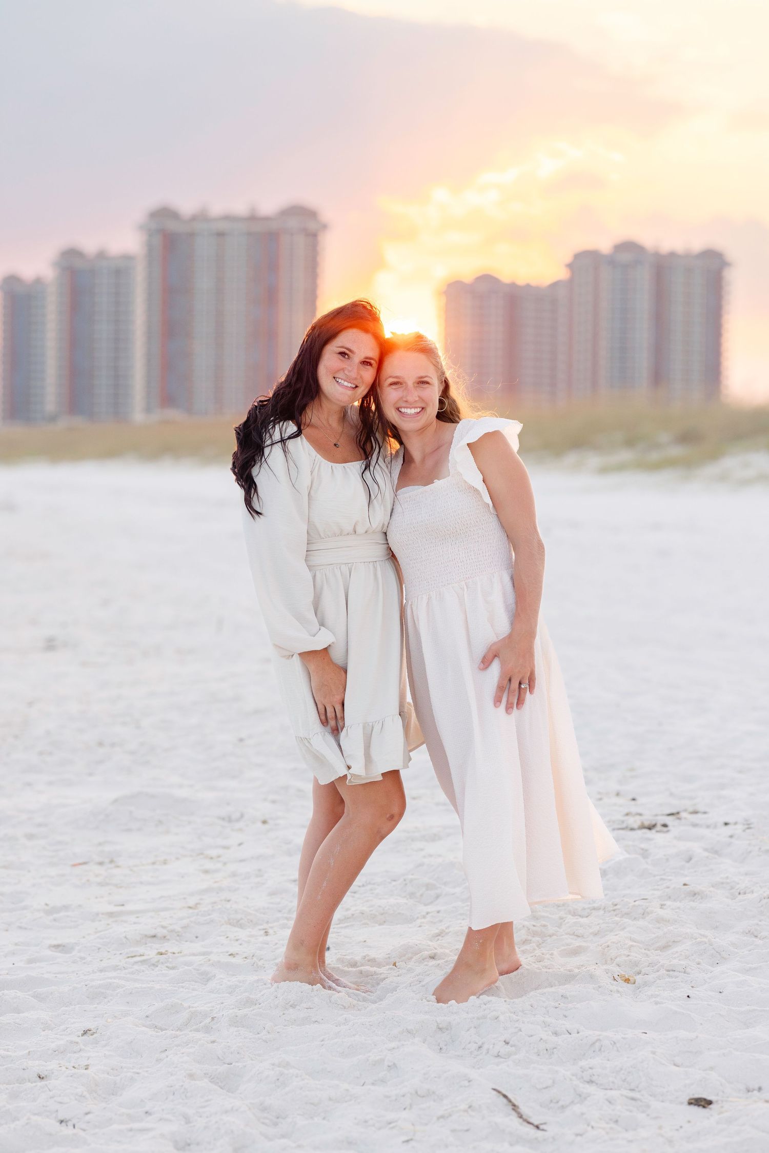 Children in white dresses playing on a sandy beach with high-rise buildings in the background at sunset.