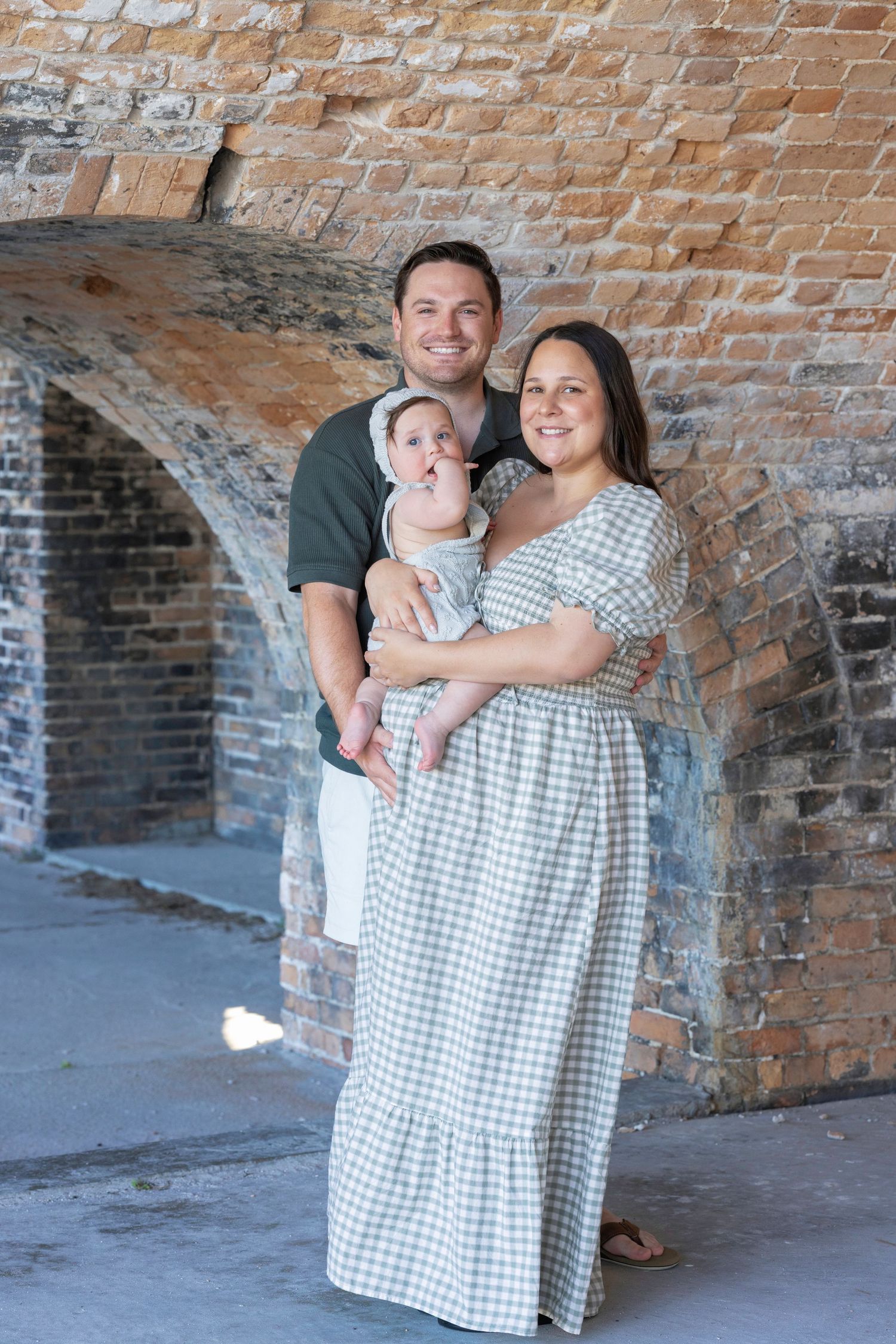 Happy family portrait with parents holding baby under brick archway outdoors.
