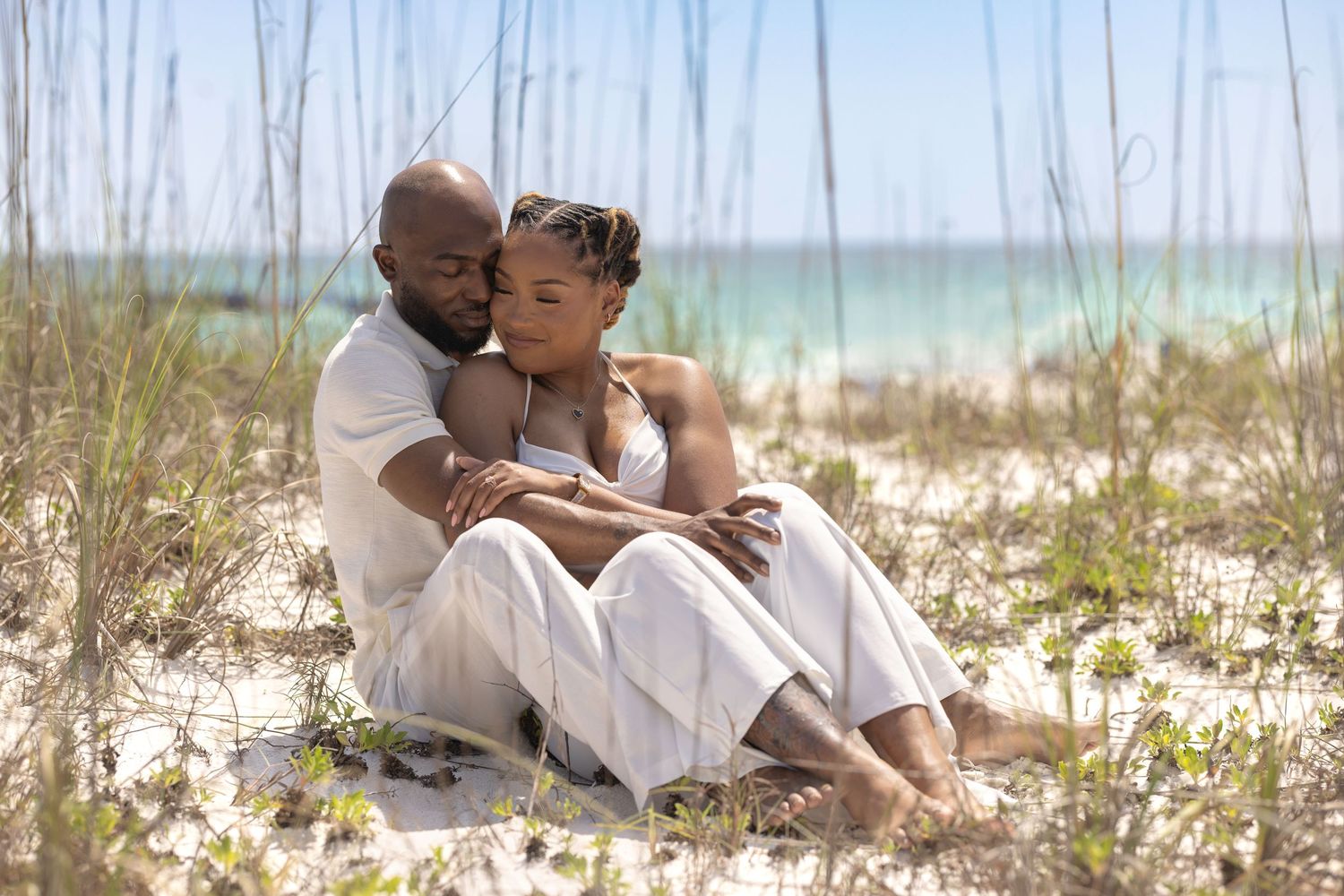 Couple sitting together on white sand beach surrounded by sea grass, wearing white outfits with turquoise ocean backdrop.