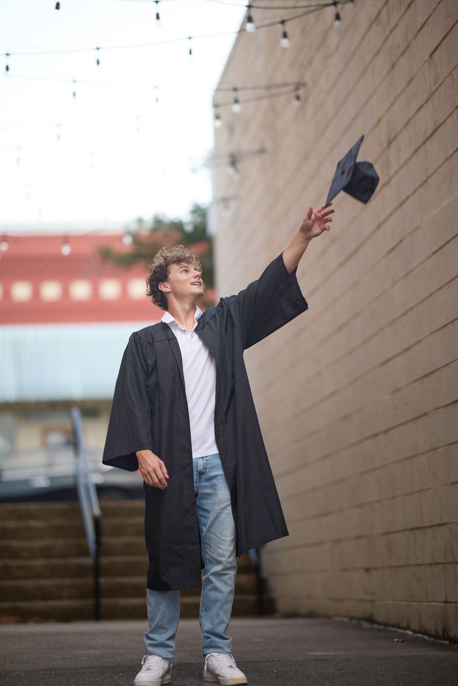 Graduate in black cap and gown tossing mortarboard into air outdoors near brick wall.